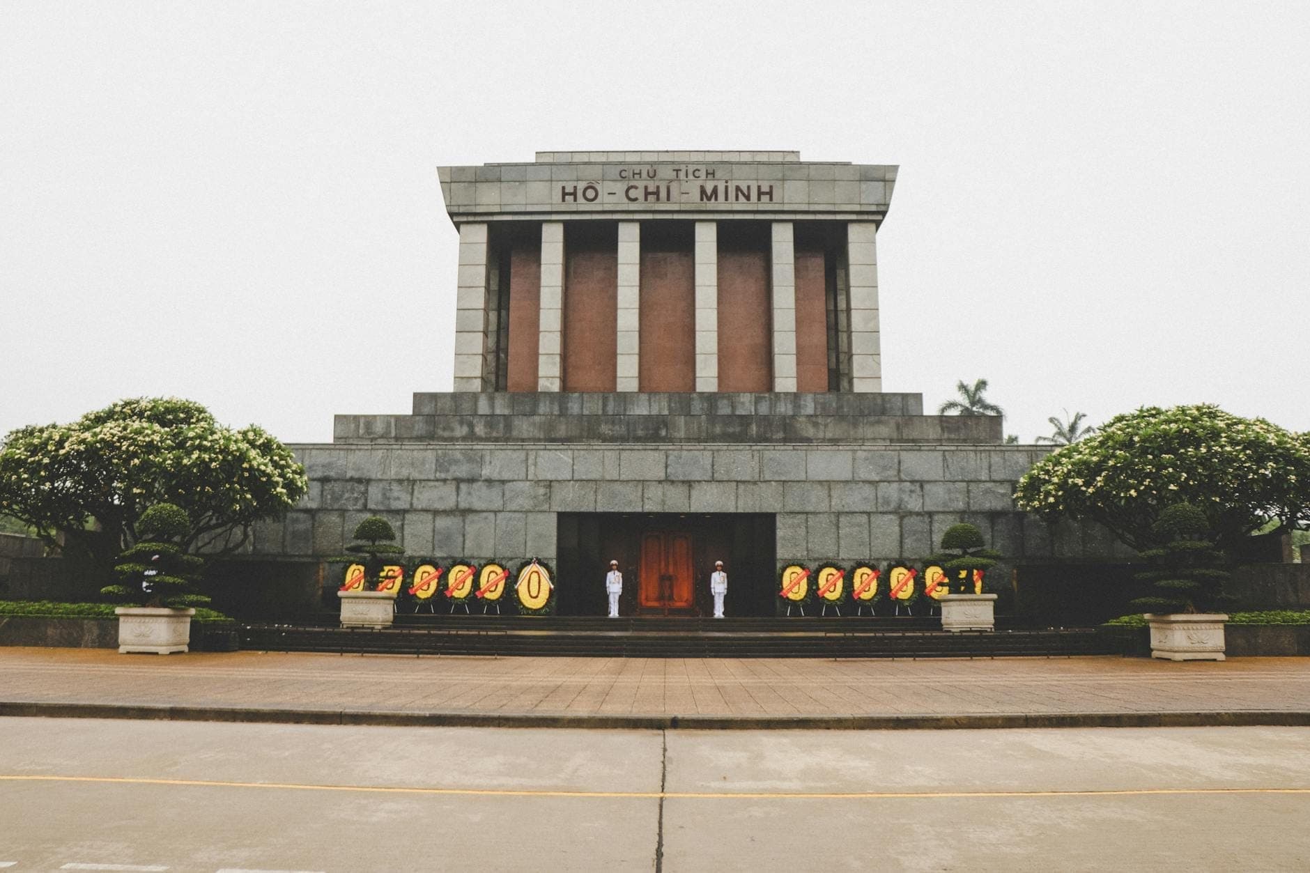 Harapang tingin ng Mausoleo ni Ho Chi Minh sa Ba Đình na may mga seremonyal na bantay at mga bulaklak, napapalibutan ng mga punong maayos ang pagkaputol at ulap sa langit.