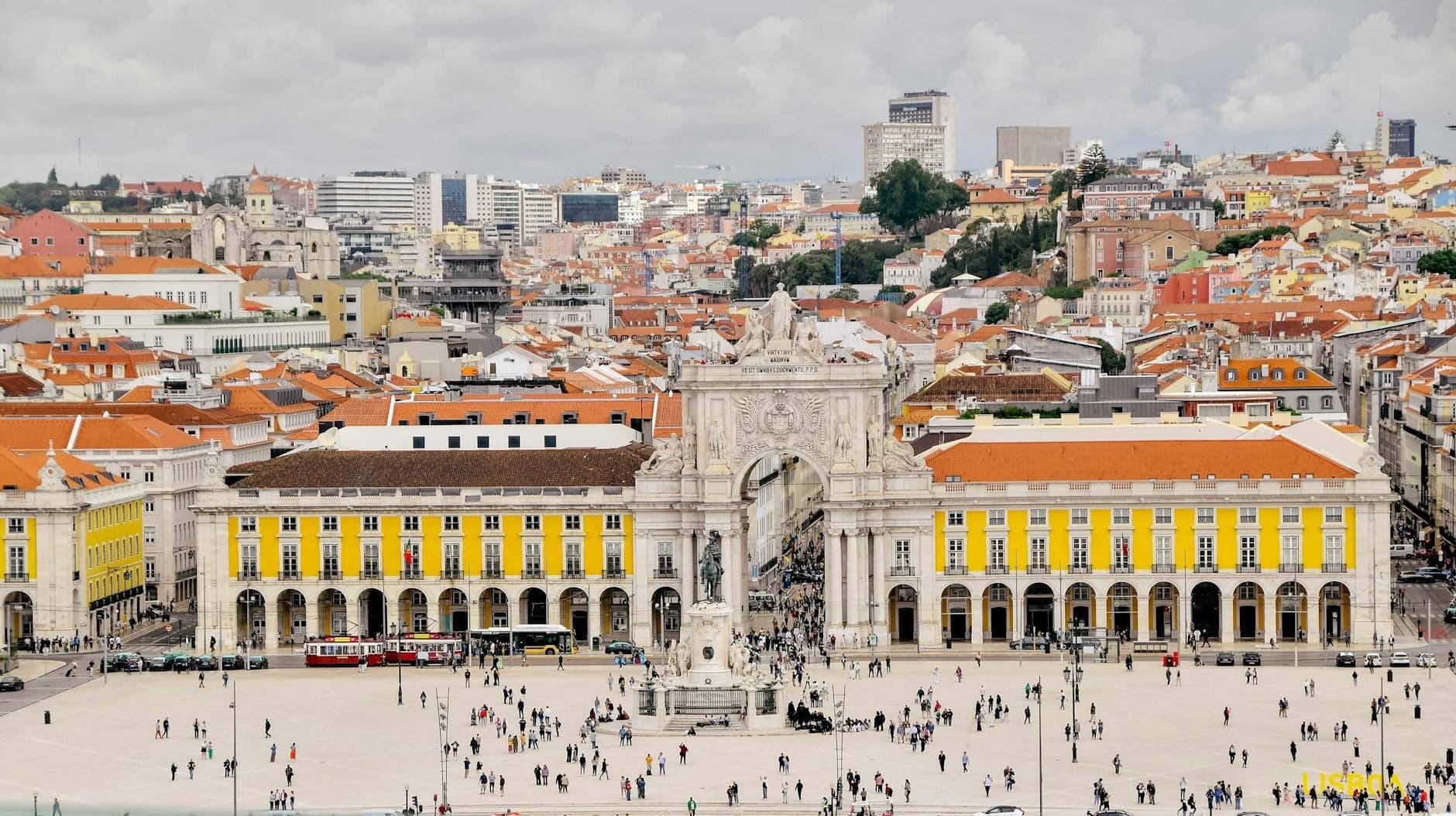 Isang malawak na panoramikong tanawin ng Praça do Comércio ng Lisbon sa Baixa, na may mga dilaw na gusali, ang Arco da Rua Augusta, at masayang buhay ng lungsod laban sa backdrop ng mga burol.