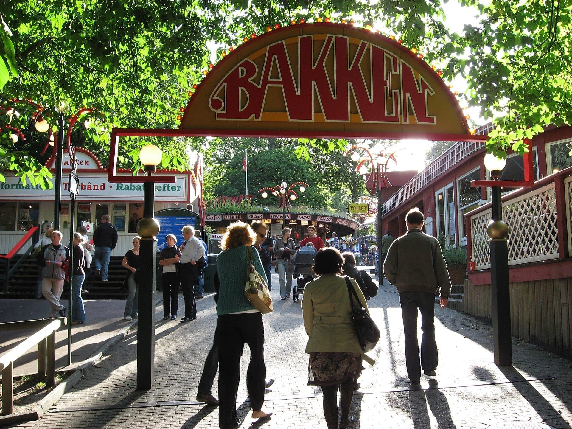 Besucher gehen unter dem ikonischen gelb-roten Eingangsschild von Bakken hindurch, umgeben von belaubten Bäumen und klassischen Vergnügungsparkgebäuden bei Tageslicht.