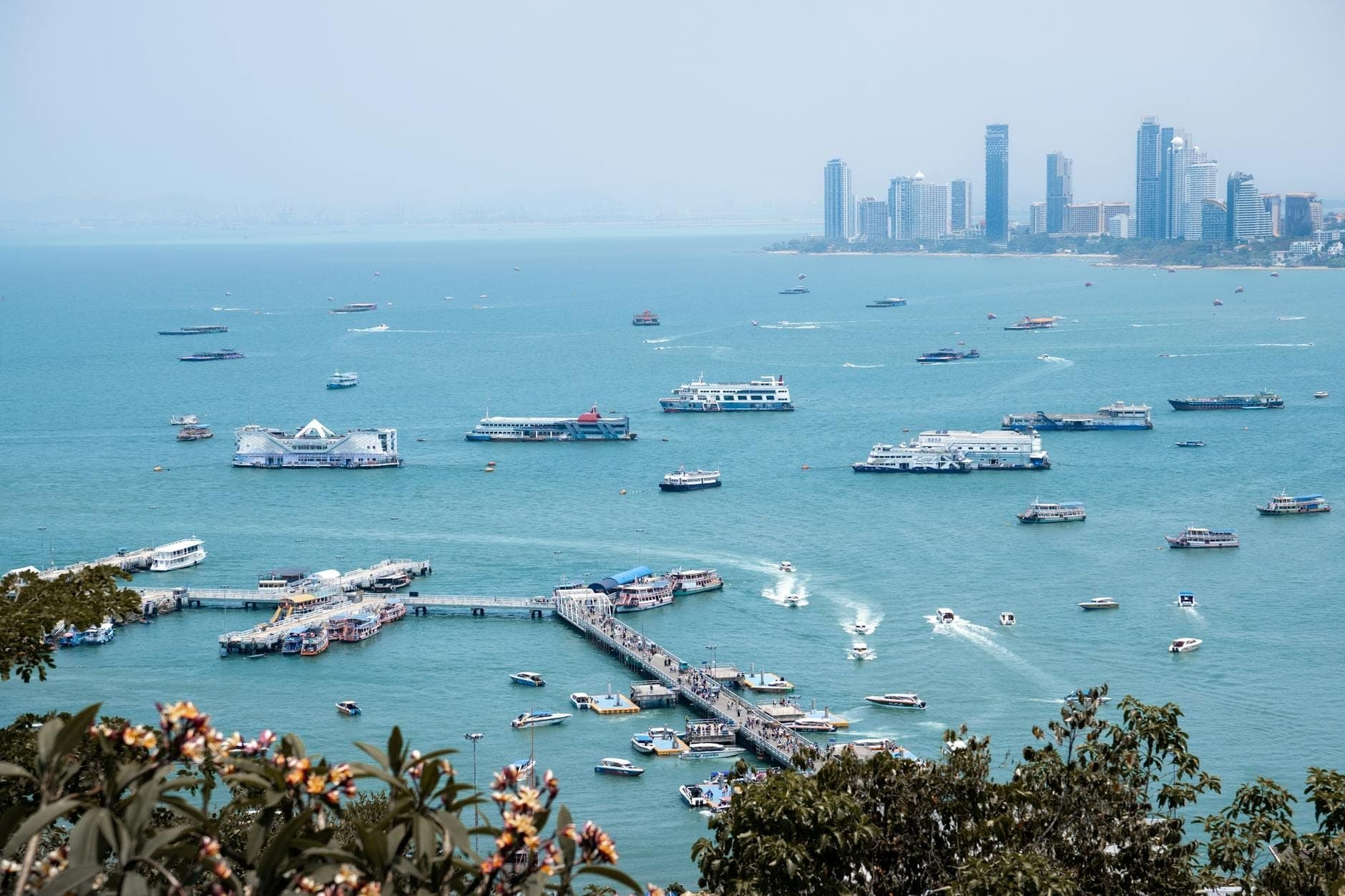 Wide view of Bali Hai Pier in Pattaya with many ferries and boats docked, turquoise sea, flowers in the foreground, and city skyline in the background.