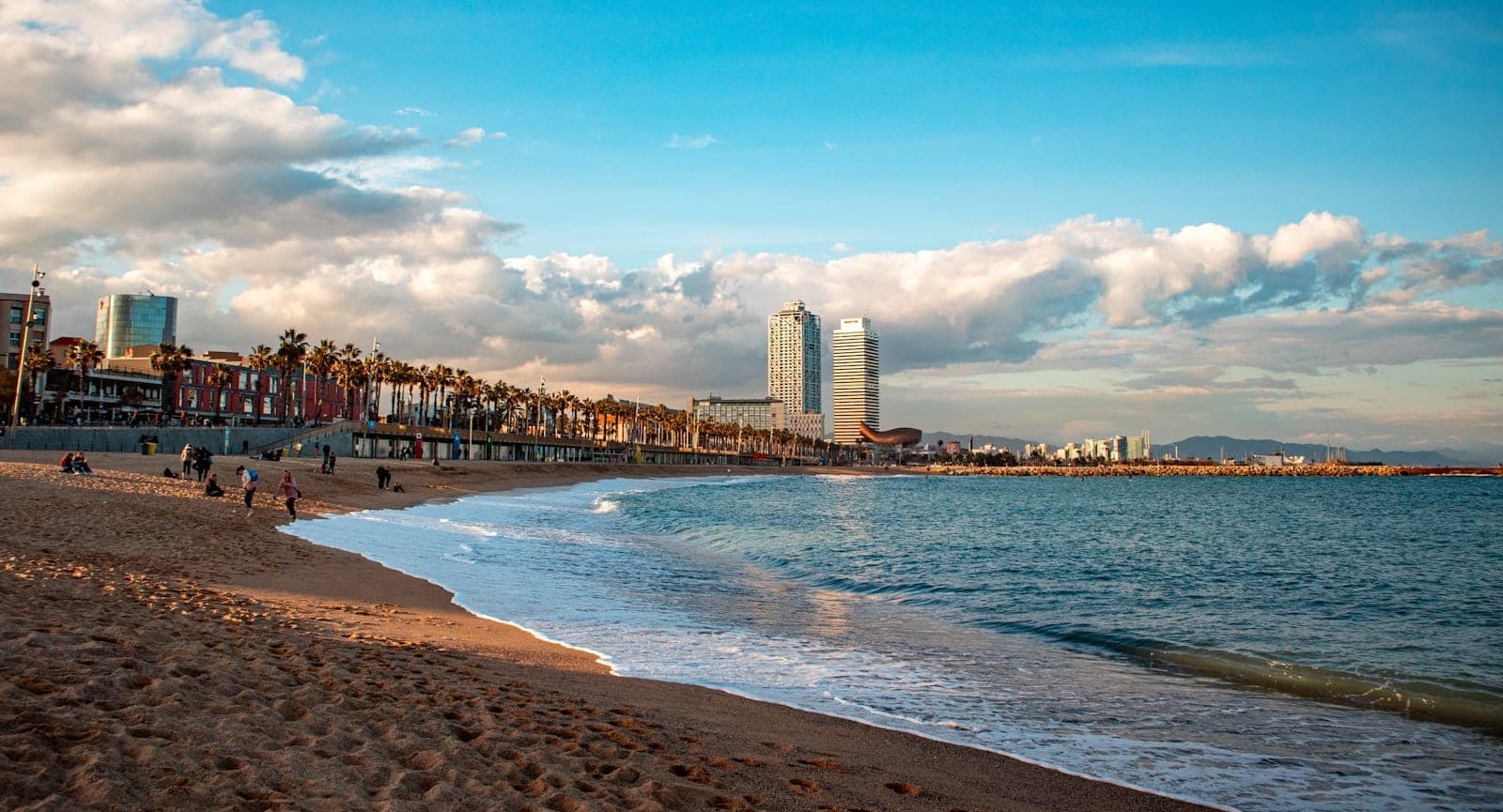 Bãi biển Barcelona rộng lớn với cát vàng và sóng nhẹ, đường chân trời thành phố với hai tòa tháp biểu tượng và hàng cọ, dưới bầu trời xanh ngắt với những đám mây trắng bồng bềnh.