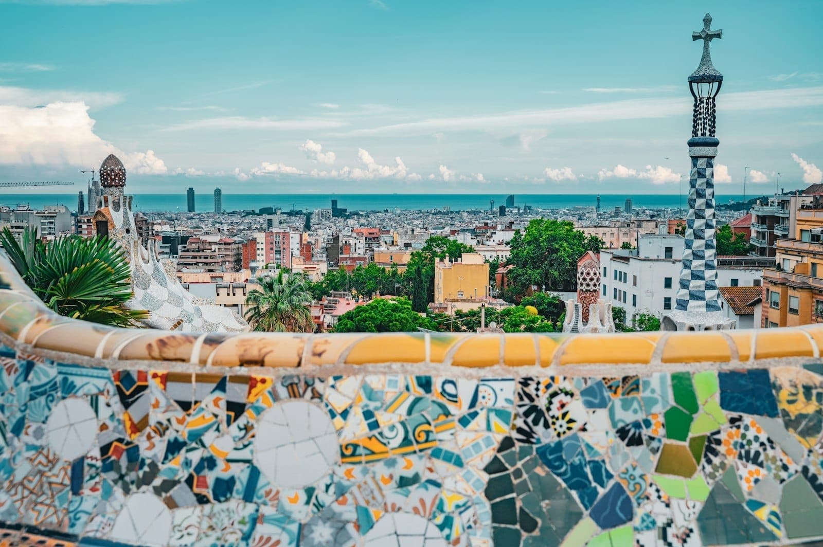 Toàn cảnh Barcelona nhìn từ Công viên Güell với ghế khảm mosaic rực rỡ ở tiền cảnh, những ngọn tháp độc đáo, cây xanh tươi tốt và thành phố trải dài ra tận biển dưới bầu trời xanh trong.