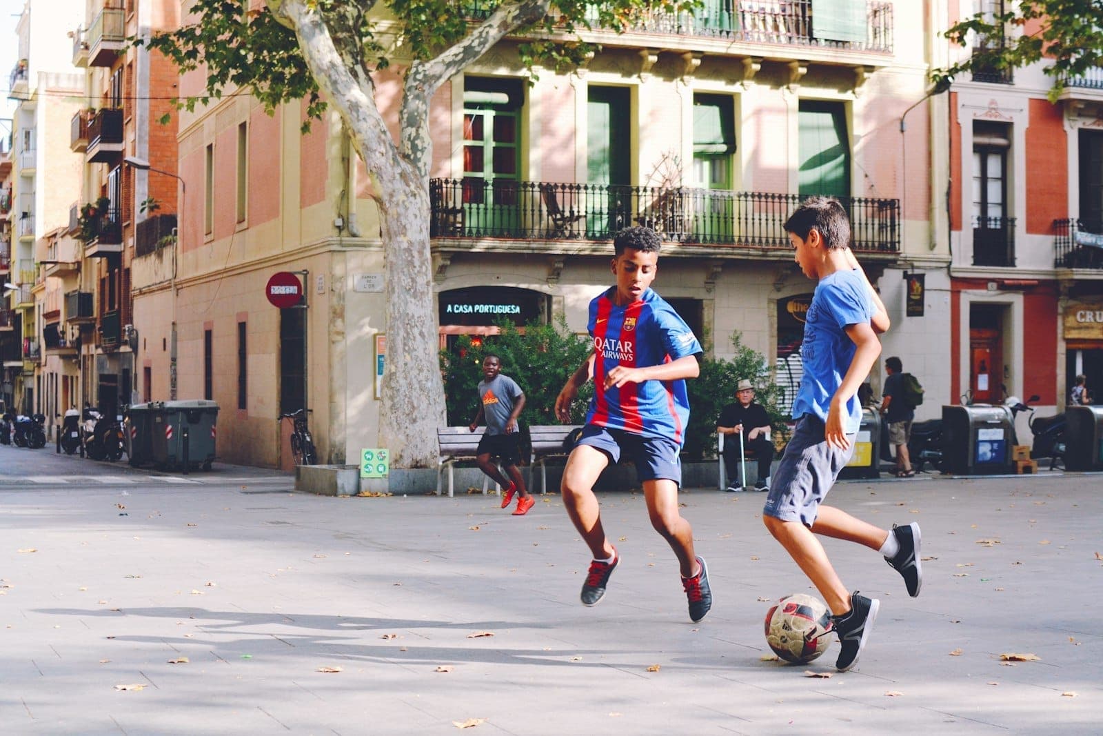 Hai đứa trẻ đang đá bóng tại một quảng trường Barcelona ngập nắng, xung quanh là những tòa nhà đầy màu sắc và cây xanh — khung cảnh tràn đầy năng lượng của chuyến du lịch gia đình.