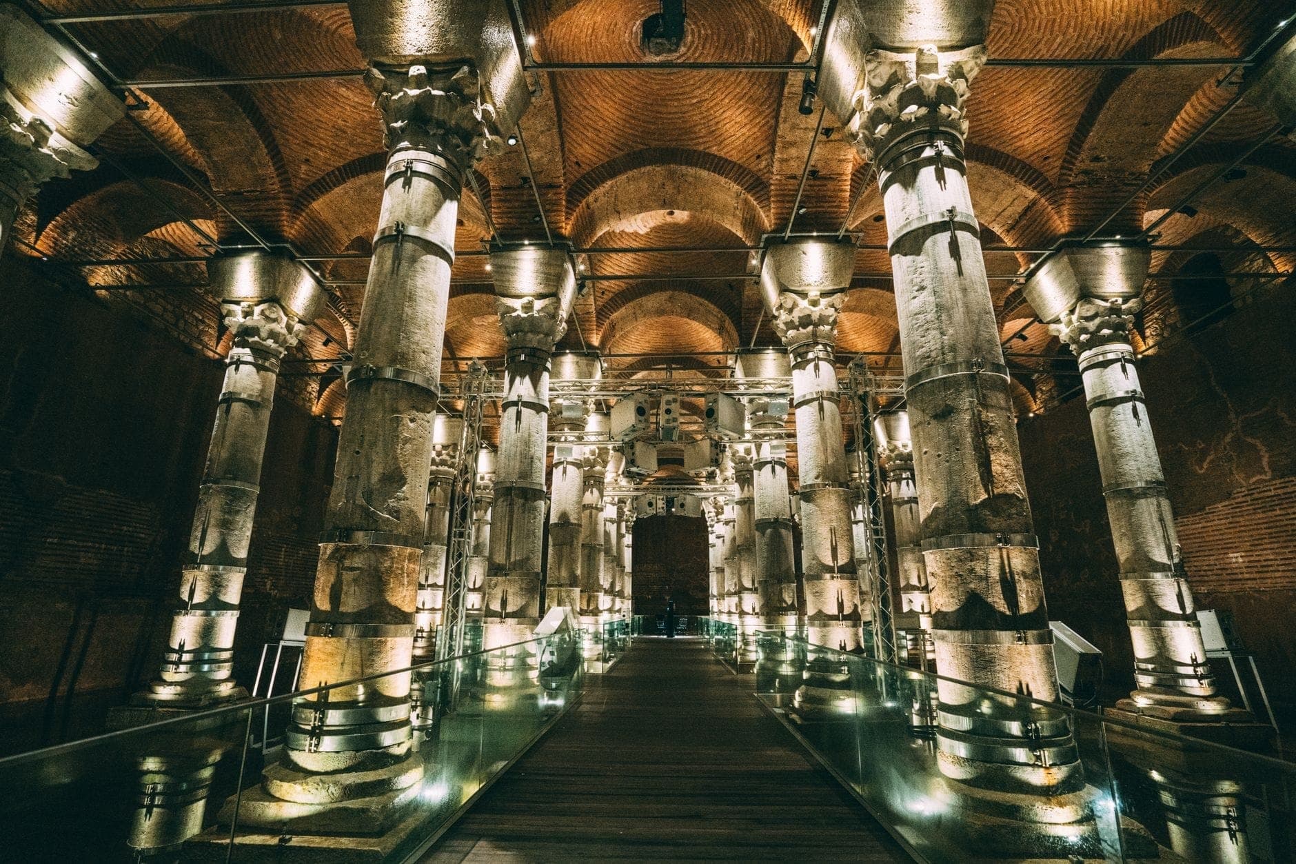 Weitwinkelansicht des Inneren der Basilika-Zisterne mit beleuchteten antiken Säulen, gewölbten Backsteindecken und einem Mittelsteg über dem spiegelglatten Wasser.