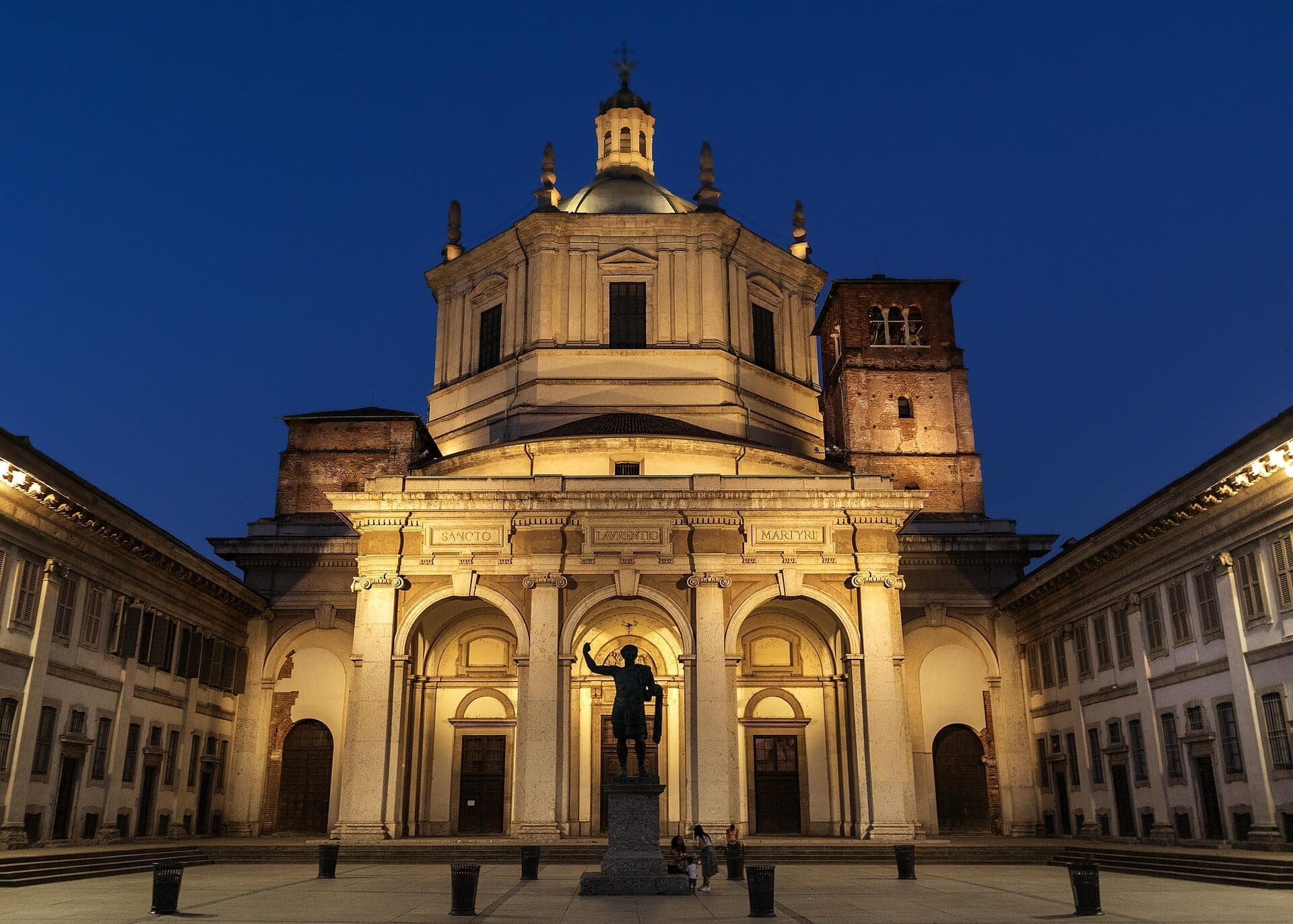 Basilica di San Lorenzo Maggiore illuminated at dusk, showing Roman columns, the central dome, and a statue in the foreground.