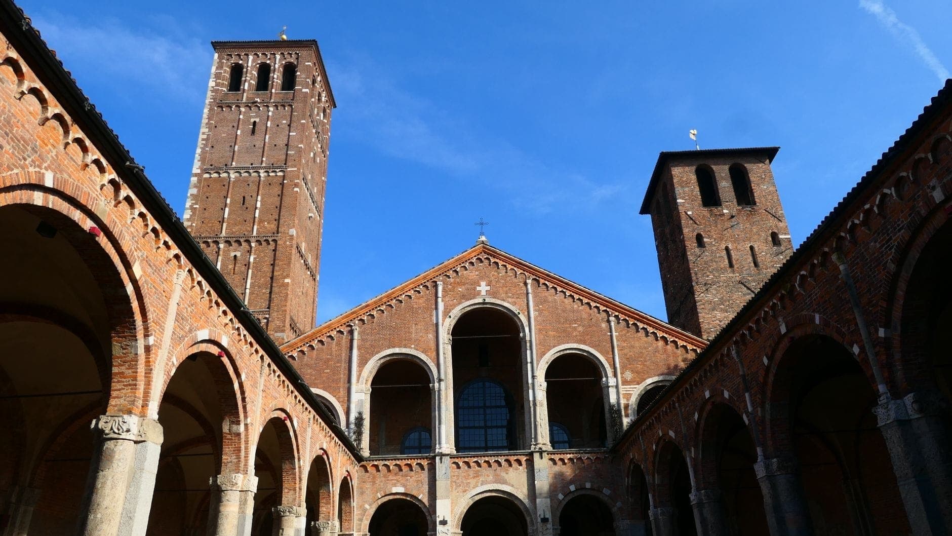 View of the Basilica di Sant'Ambrogio courtyard and facade with two Romanesque towers and arched entry, under a clear blue sky in Milan.