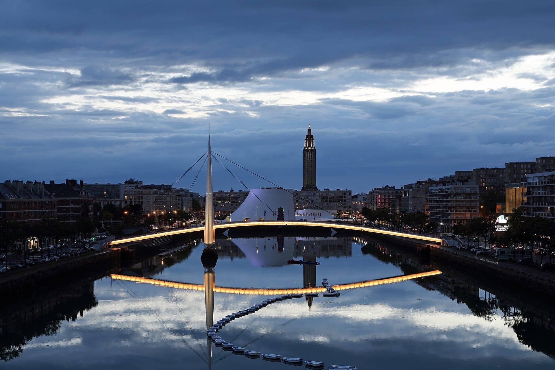 Cảnh buổi tối tại Bassin du Commerce ở Le Havre, nổi bật với những cây cầu hiện đại, các tòa nhà ven sông và hình phản chiếu của tháp dưới bầu trời mây ấn tượng.