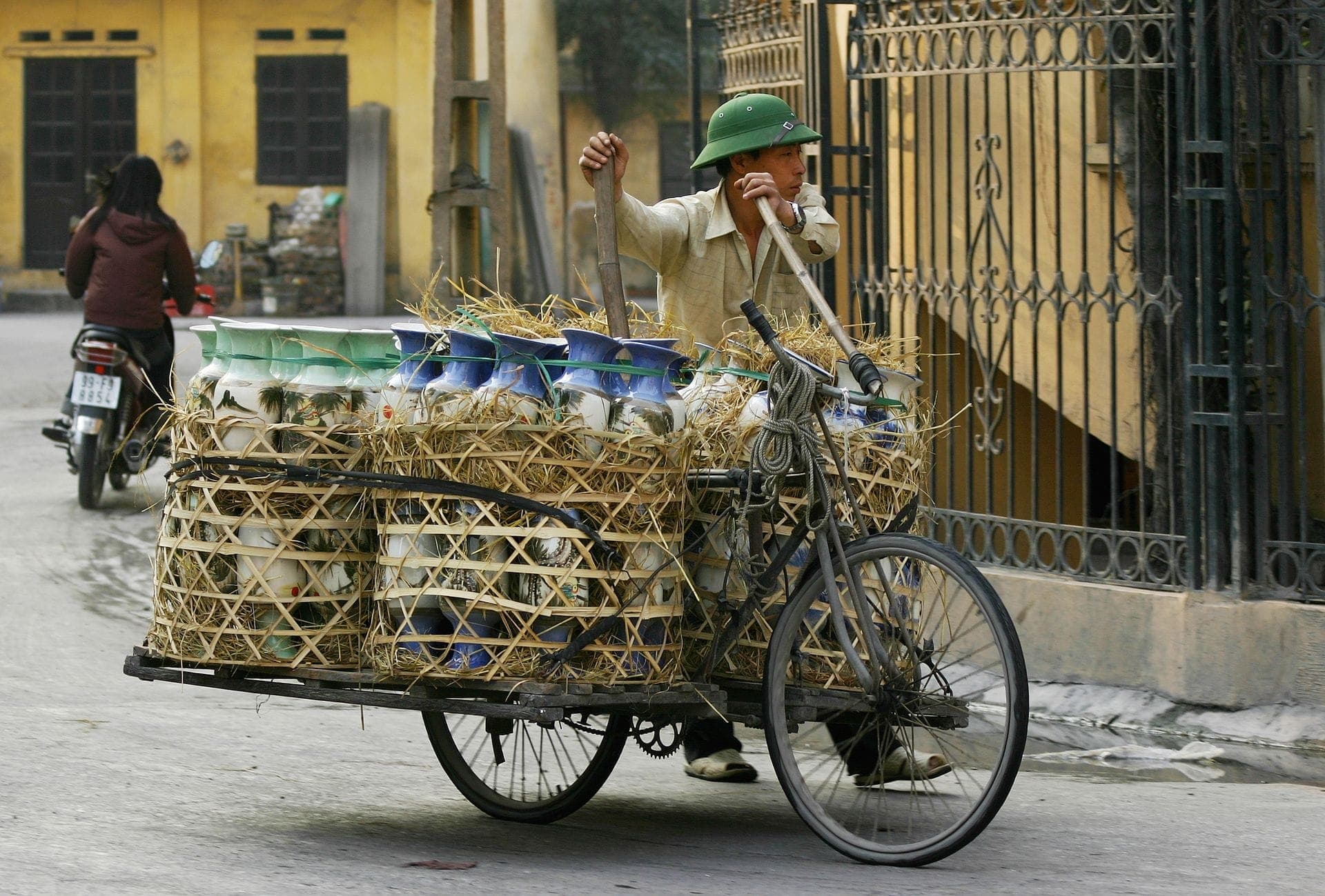 Một người đàn ông vận chuyển những chiếc bình gốm lớn được vẽ trang trí bằng xe đạp thồ qua các con phố của làng gốm Bát Tràng, với những ngôi nhà màu vàng ở phía sau.