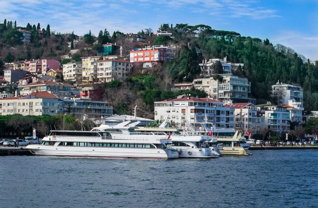 Bunte Wohngebäude und Yachten säumen das Ufer der Bebek Uferpromenade, im Hintergrund üppige grüne Hügel am Bosporus in Istanbul.