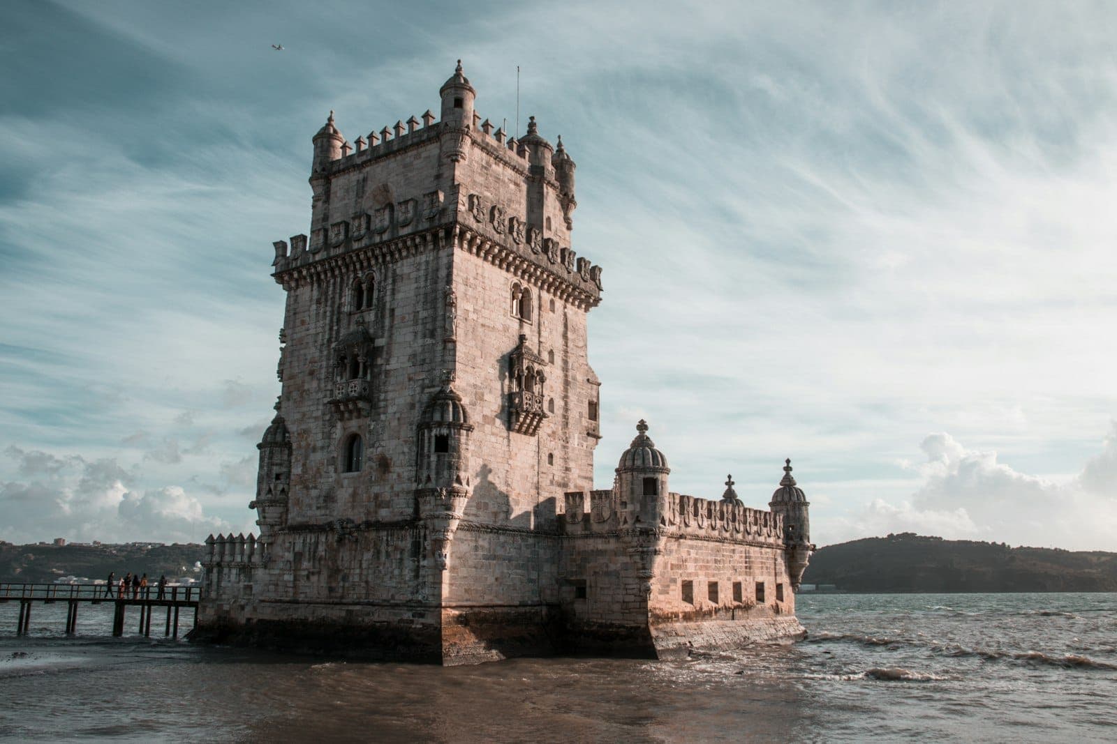 Ang Torre de Belém na nakatayo sa tabi ng ilog sa ilalim ng dramatikong langit, na may masalimuot na arkitekturang Manueline at tubig sa harapan.