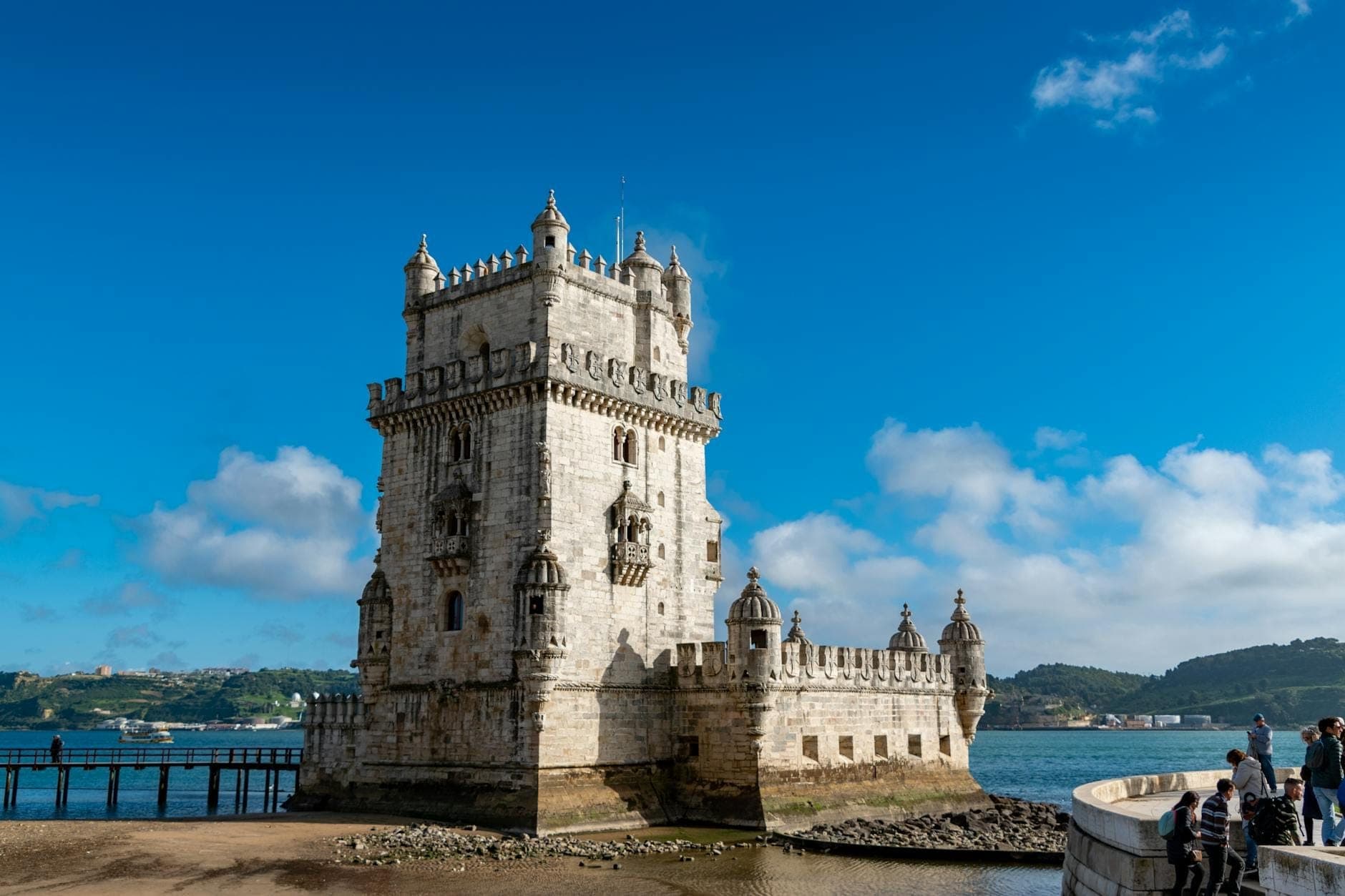 Tháp Belém sừng sững bên sông Tagus dưới bầu trời xanh với những đám mây trắng, du khách dạo bước gần đó và thành phố Lisbon hiện ra phía sau.