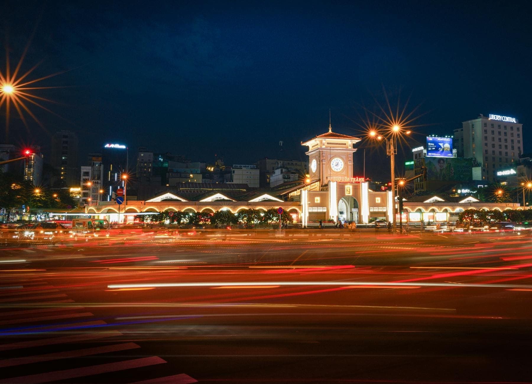 Chợ Bến Thành về đêm với tháp đồng hồ sáng rực, dòng xe tạo vệt sáng trên đường và các tòa nhà thành phố phía sau.