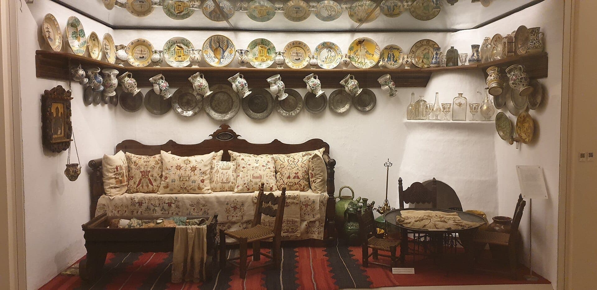 Traditional Greek sitting room display at Benaki Museum, featuring embroidered cushions, wooden furniture, and shelves of decorative ceramics against whitewashed walls.