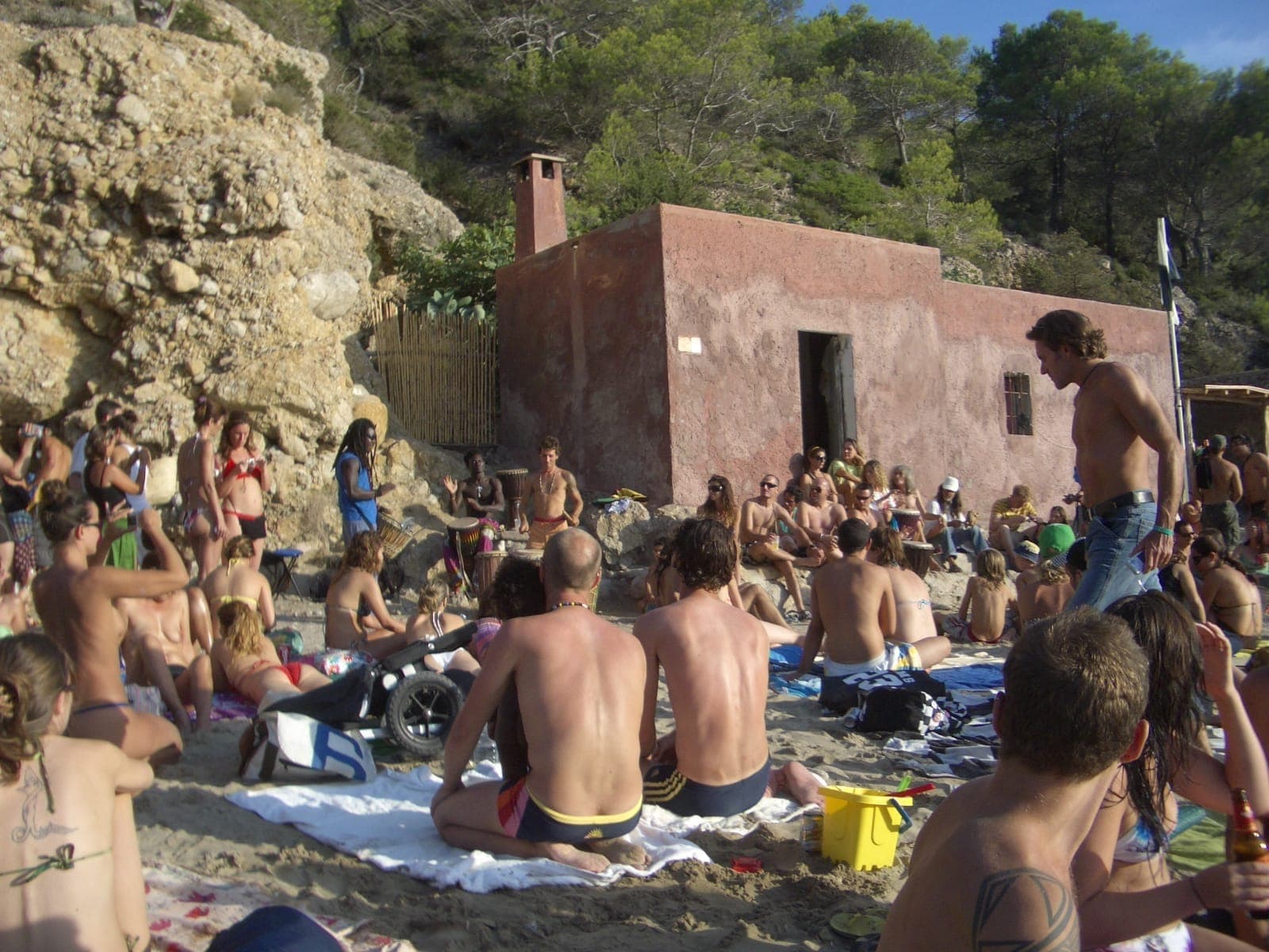 Visitors gather on the sandy shore of Benirràs Beach, Ibiza, lounging near a rustic building surrounded by rocky cliffs and pine trees.