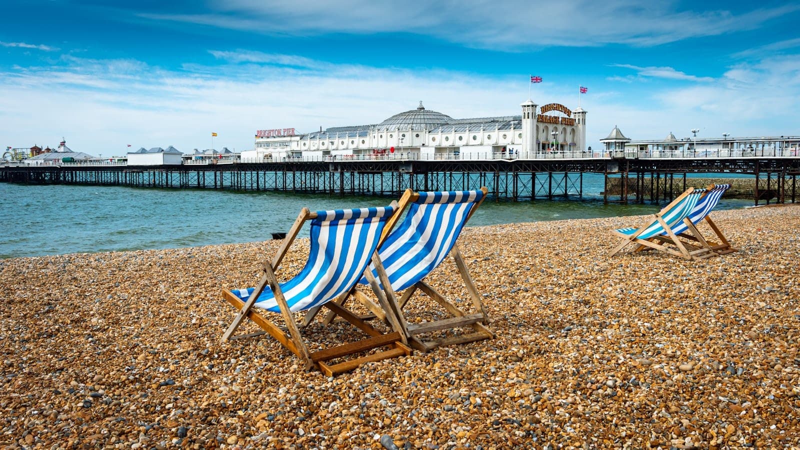 Ghế bãi biển sọc đặt trên bãi sỏi Brighton với cầu tàu Brighton và bầu trời xanh phía sau, không khí biển cổ điển đặc trưng.