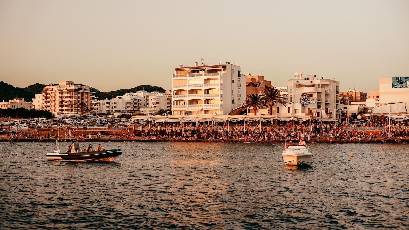 Beachfront scene in Ibiza with lively crowds, numerous bars and restaurants, and boats anchored just offshore at sunset.