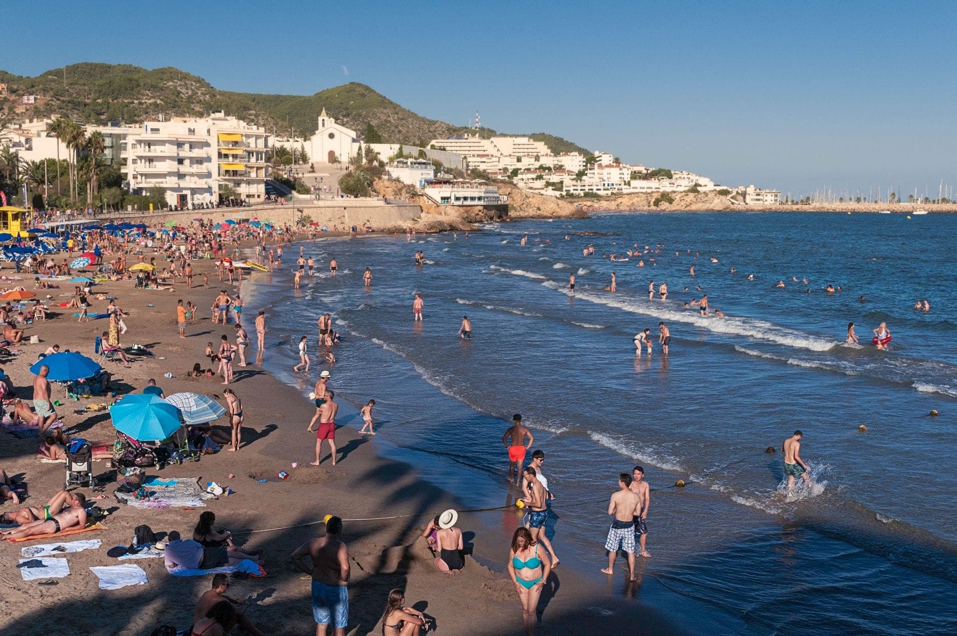 Busy beach scene with families and children on the sand and in the shallow water, buildings of Santa Eulalia in the background.