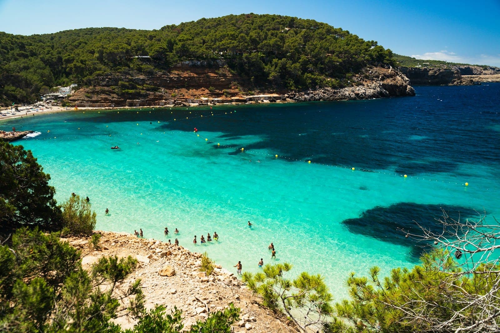 Scenic Ibiza beach with turquoise water, swimmers, and lush green hills in the background under a bright sunny sky.
