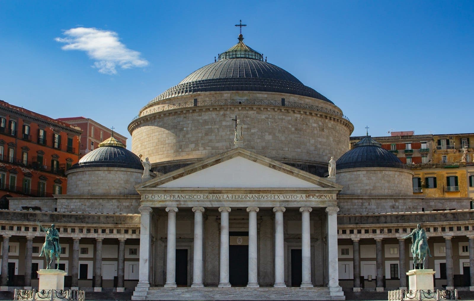 Toàn cảnh mặt tiền nhà thờ San Francesco di Paola với mái vòm lớn, bầu trời xanh, hai bên là các bức tượng và những tòa nhà đầy màu sắc tại Naples, Ý.
