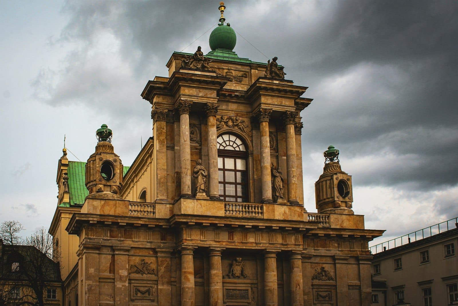 Barocke Kirchenfassade in Warschau mit verzierten Säulen und Statuen, dramatische Beleuchtung unter bewölktem Himmel – ein Blick auf historische Architektur.