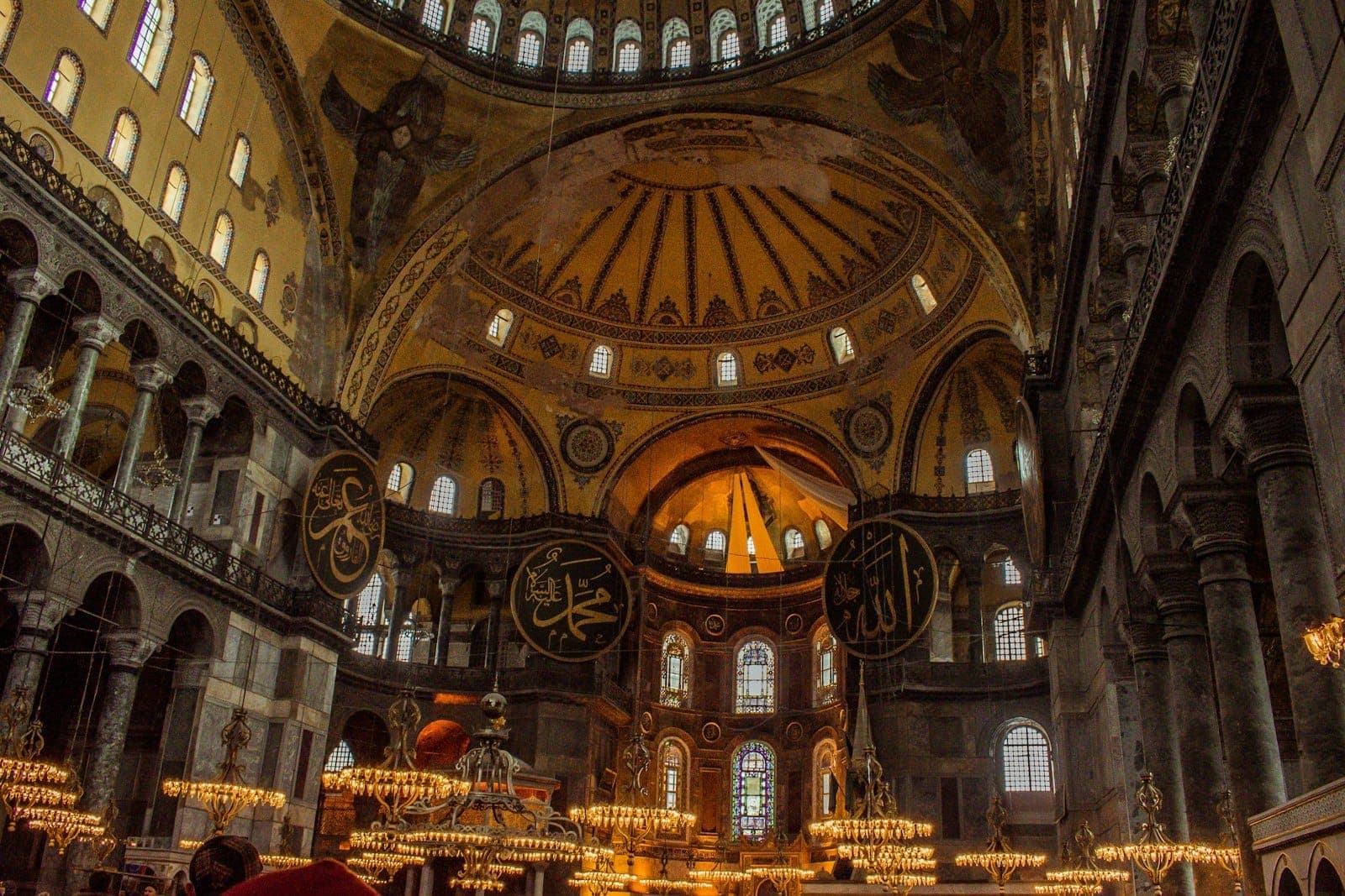 Vista amplia de la gran cúpula y las galerías superiores de Hagia Sophia, con grandes medallones caligráficos y detalles arquitectónicos históricos dorados.