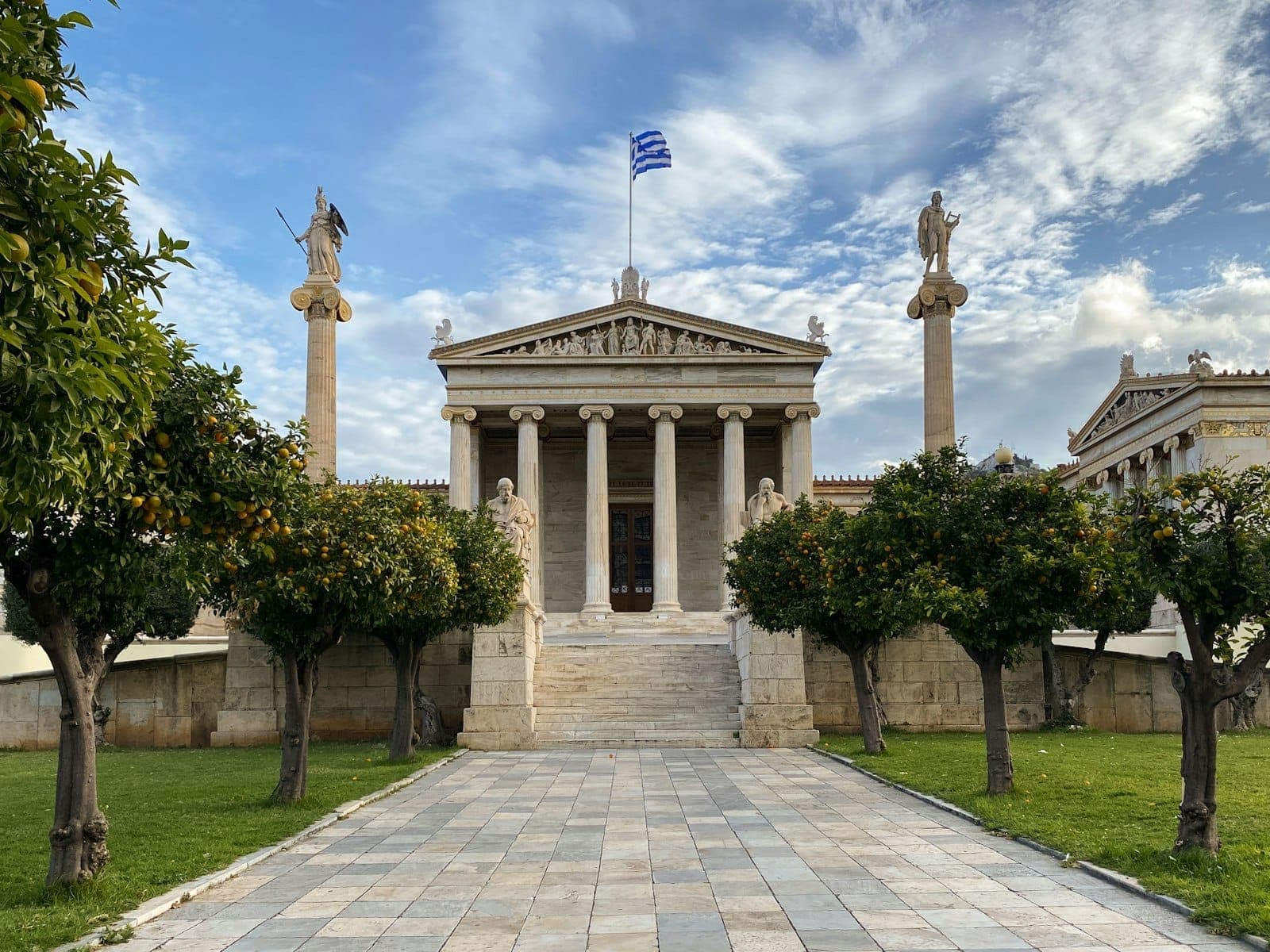 Grand neoclassical facade of the National Archaeological Museum of Athens, lined with columns and statues, under a blue sky with Greek flag flying above.
