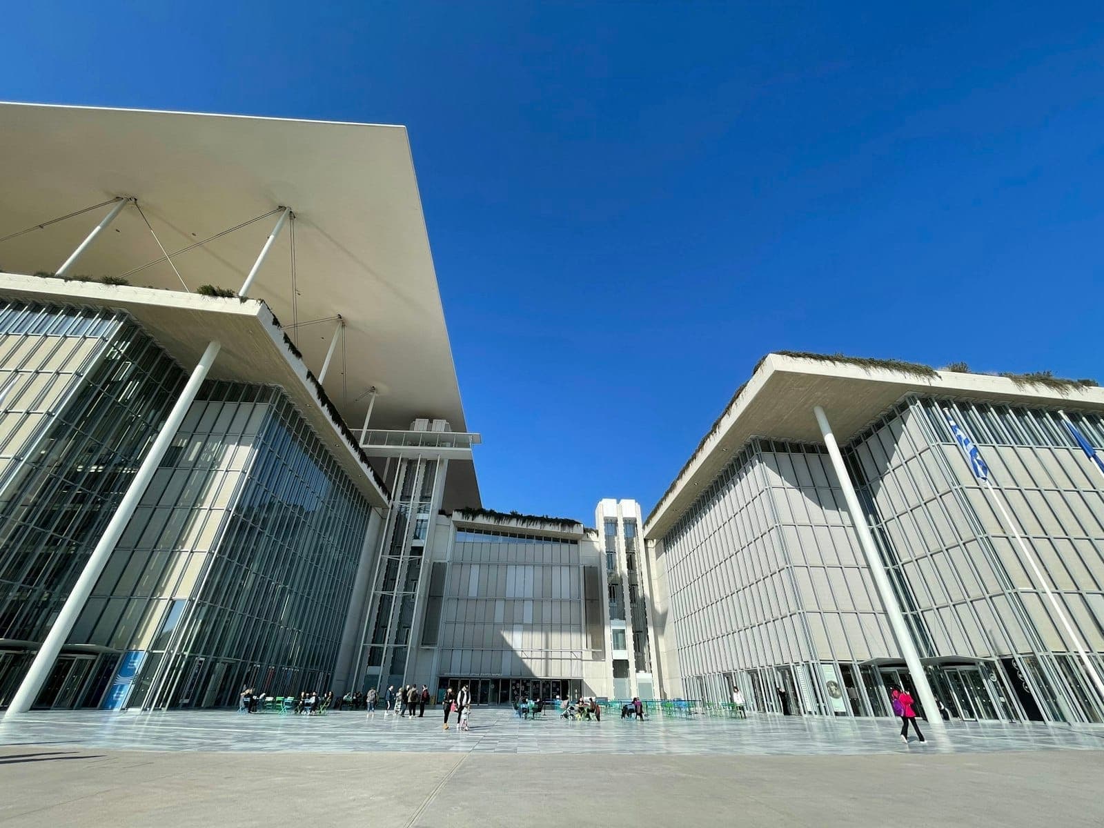 Modernong Stavros Niarchos Foundation Cultural Center na may salaming harapan sa Athens, na may asul na langit at mga tao sa plaza sa labas