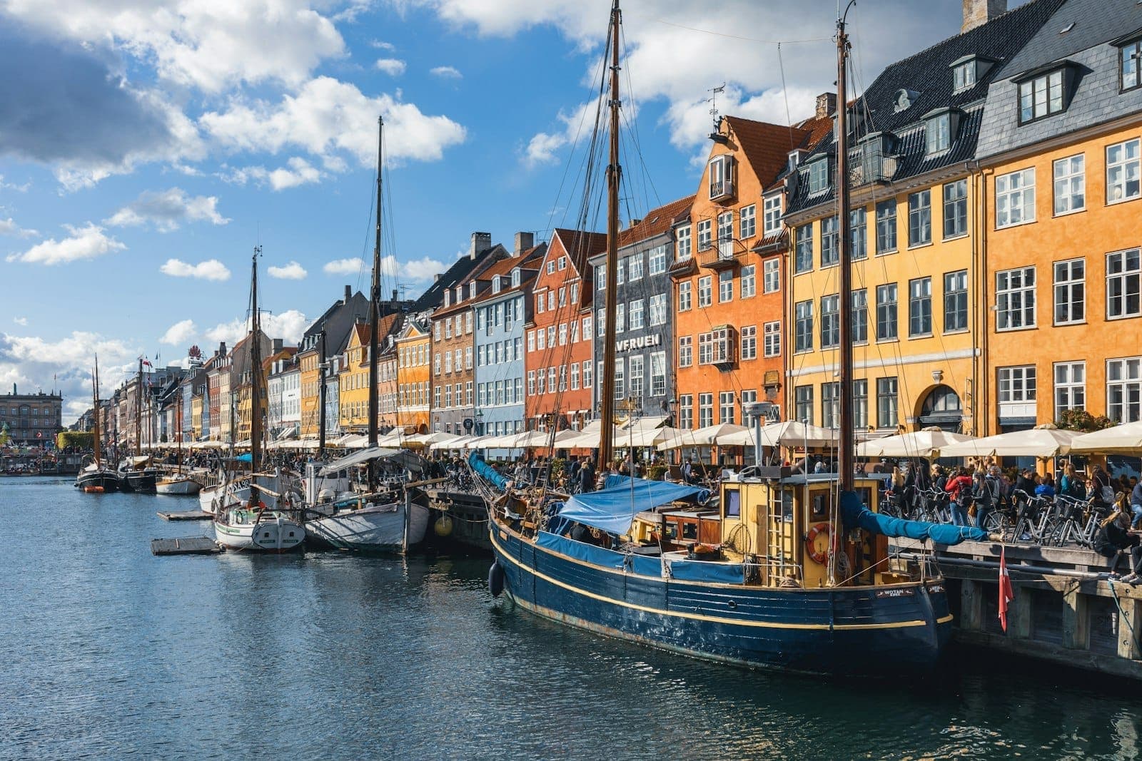 Blick auf die bunten historischen Häuser und festgemachten Boote am Nyhavn-Kanal in Kopenhagen an einem sonnigen Tag, mit vielen Menschen, die die Atmosphäre am Wasser genießen.