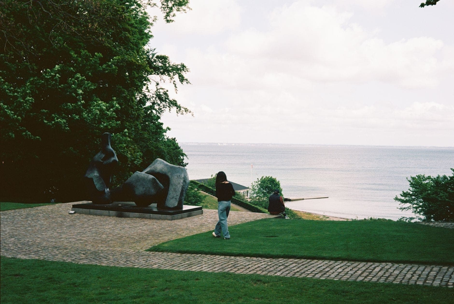 Park rzeźb z figurą nowoczesnej sztuki, ścieżką, bujnym zielonym trawnikiem i widokiem na morze, spacerujący ludzie w pobliżu Louisiana Museum.