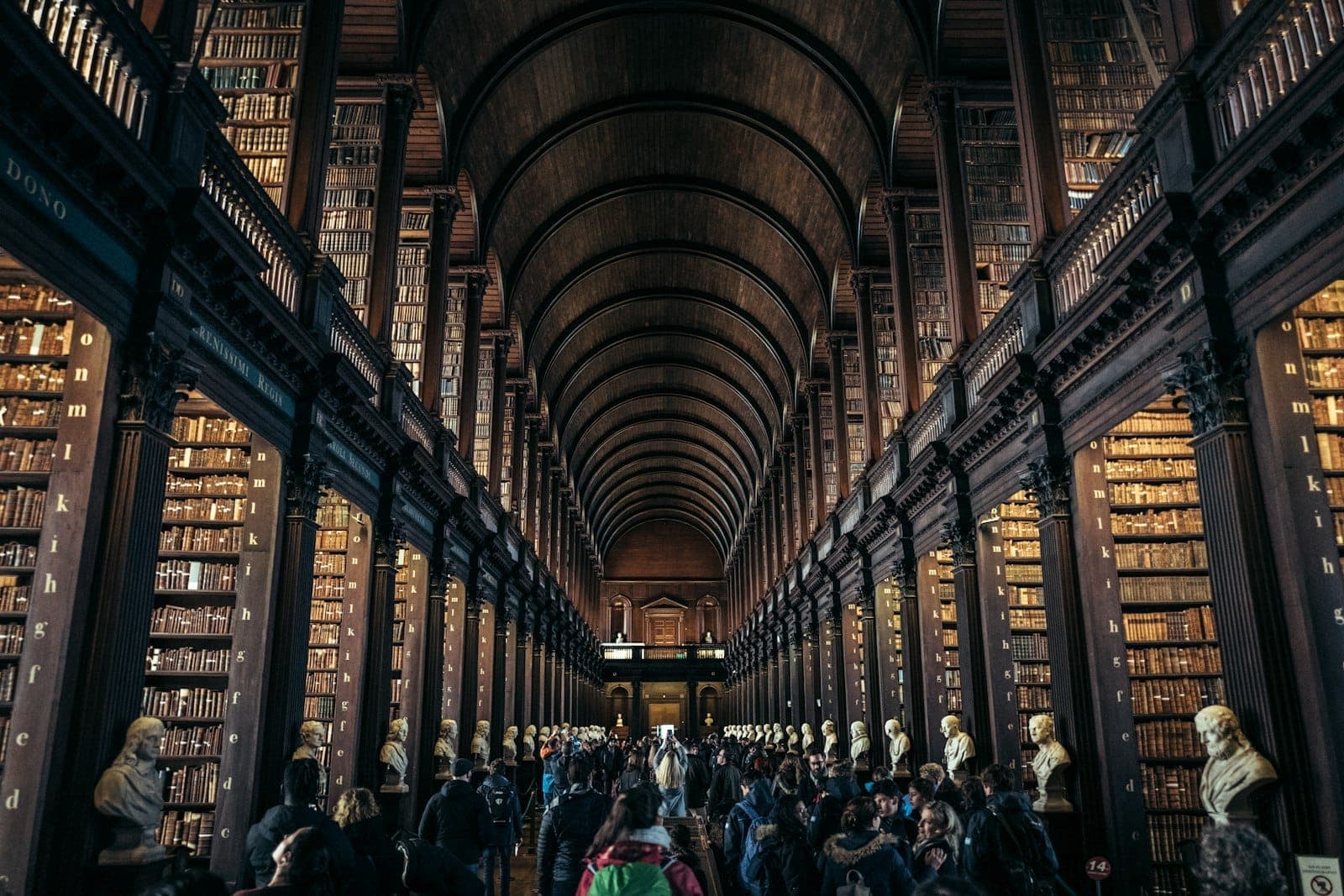 Long Room w bibliotece Trinity College w Dublinie, z wysokimi półkami na książki, łukowym drewnianym sufitem, marmurowym popiersiem i zwiedzającymi spacerującymi po wnętrzu.