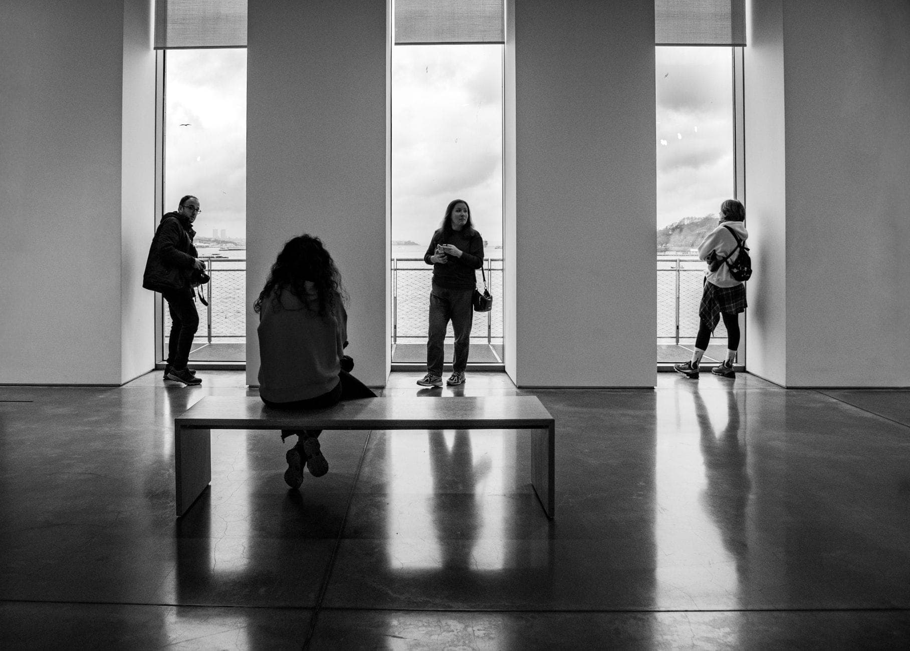 Foto en blanco y negro de personas de pie y sentadas dentro de una galería de arte moderno con grandes ventanales y vistas al Bósforo.
