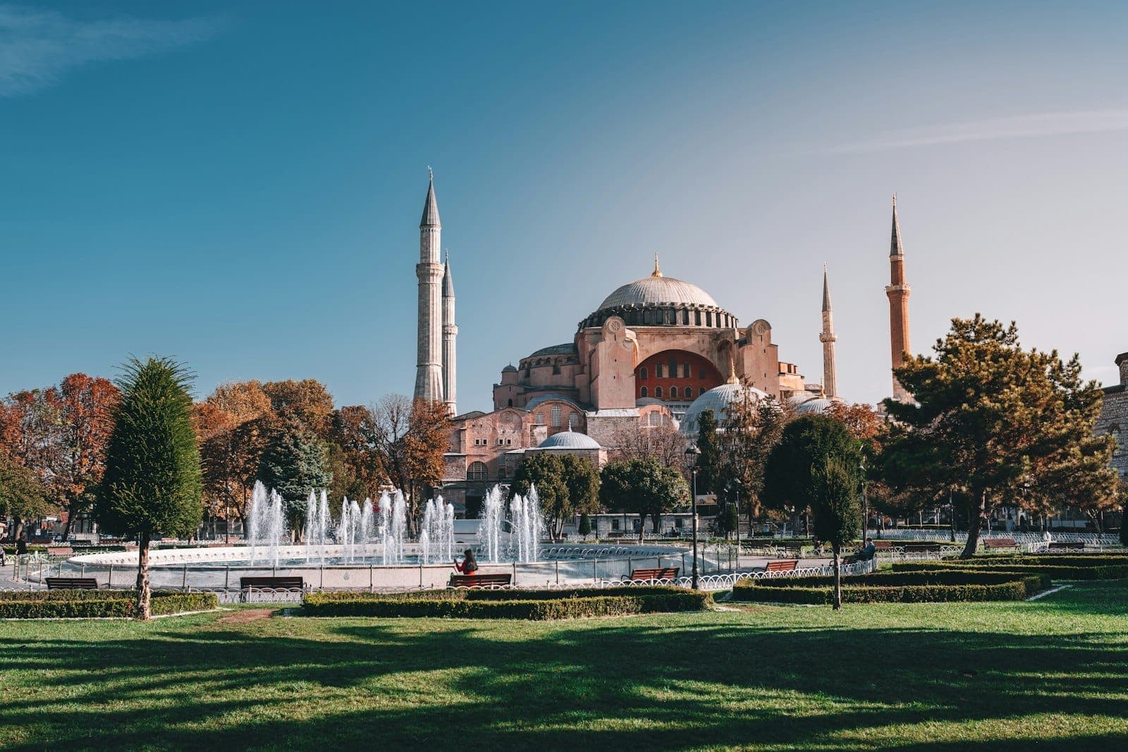 Weiter, lebendiger Blick auf die Hagia Sophia mit Minaretten, grünem Park, Bäumen und einem Brunnen an einem sonnigen Tag in Istanbul.