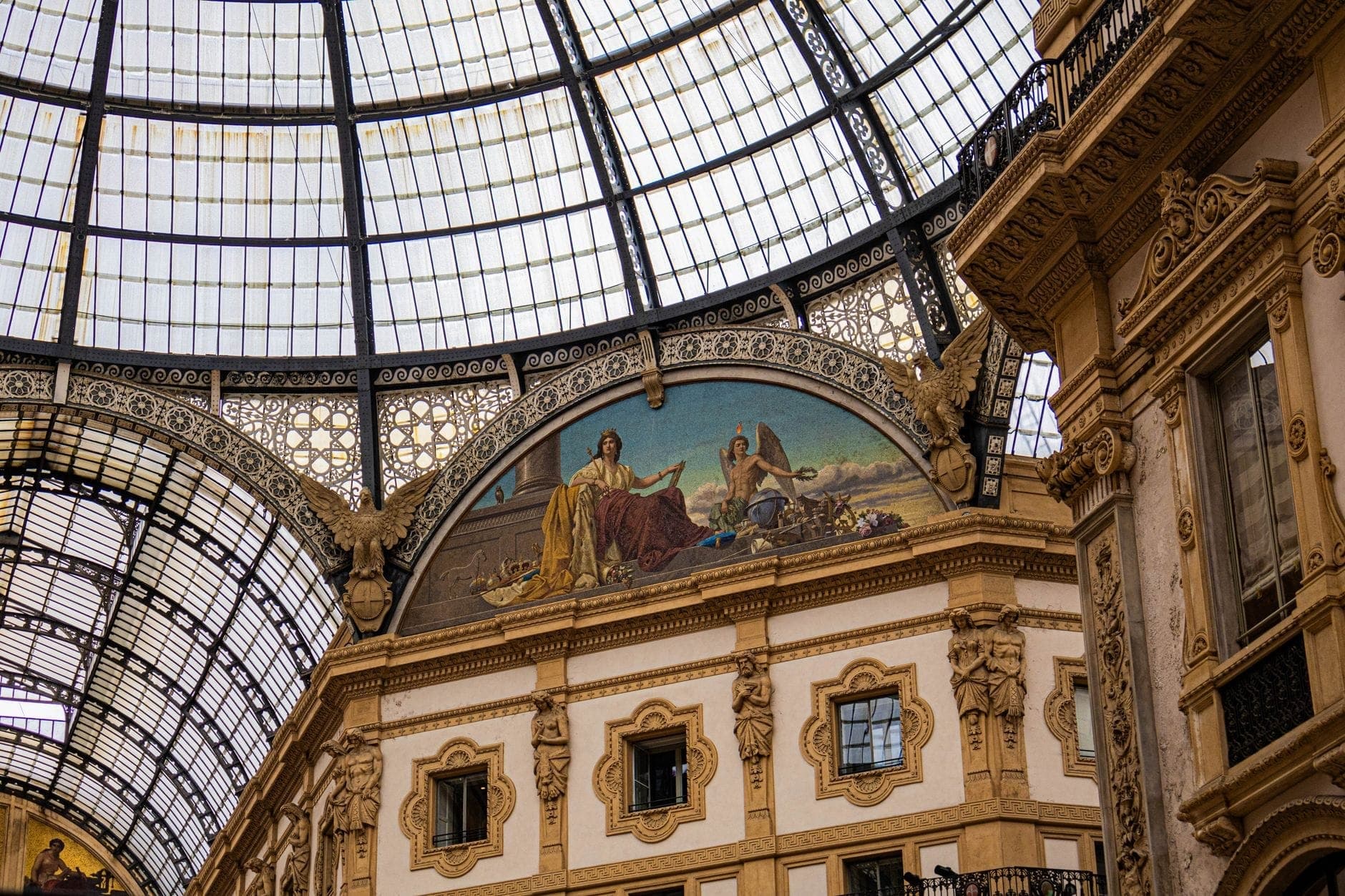Glass-domed ceiling and ornate architecture inside Galleria Vittorio Emanuele II in Milan, featuring classical artwork and detailed decorative elements.