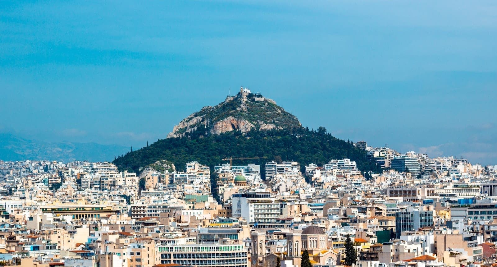 Vista panorâmica clara de Atenas com o Monte Licabeto emergindo acima dos edifícios brancos da cidade sob um céu azul.