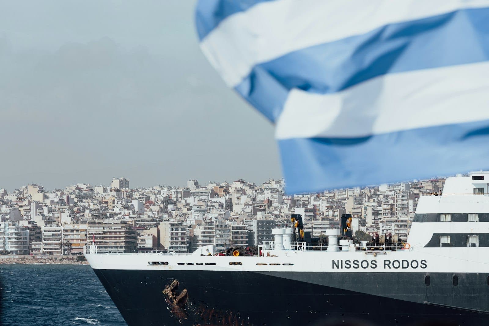 Close-up de uma grande balsa chamada Nissos Rodos no porto do Pireu, Atenas, com a bandeira grega e edifícios da cidade ao fundo.