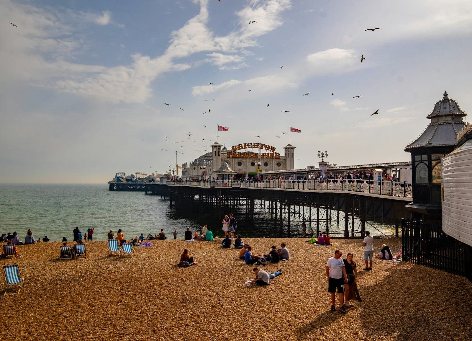 Cầu Brighton nhìn từ bãi biển sỏi vào ngày có nắng nhẹ, mọi người thư giãn tận hưởng không khí biển.