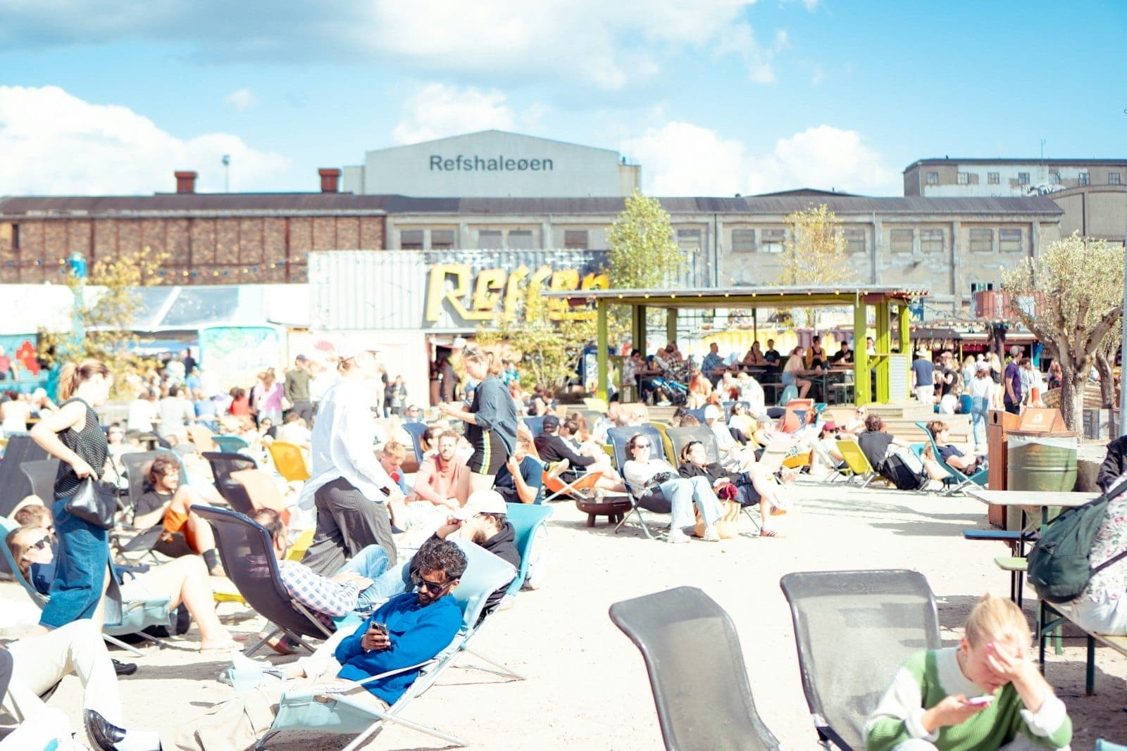 Crowd lounging outdoors in summer at Refshaleøen, Copenhagen, with festive seating and people enjoying a lively event under blue sky.