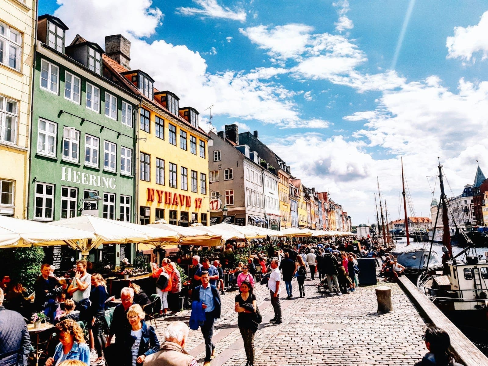 Crowds enjoy outdoor restaurants and vibrant life along Nyhavn canal on a sunny summer day in Copenhagen.