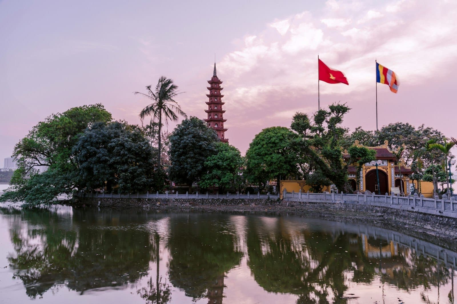 Chùa Trấn Quốc và bóng phản chiếu trên mặt hồ lúc hoàng hôn tại Hà Nội, với những lá cờ rực rỡ và cây xanh tươi tốt dưới bầu trời đầy màu sắc.
