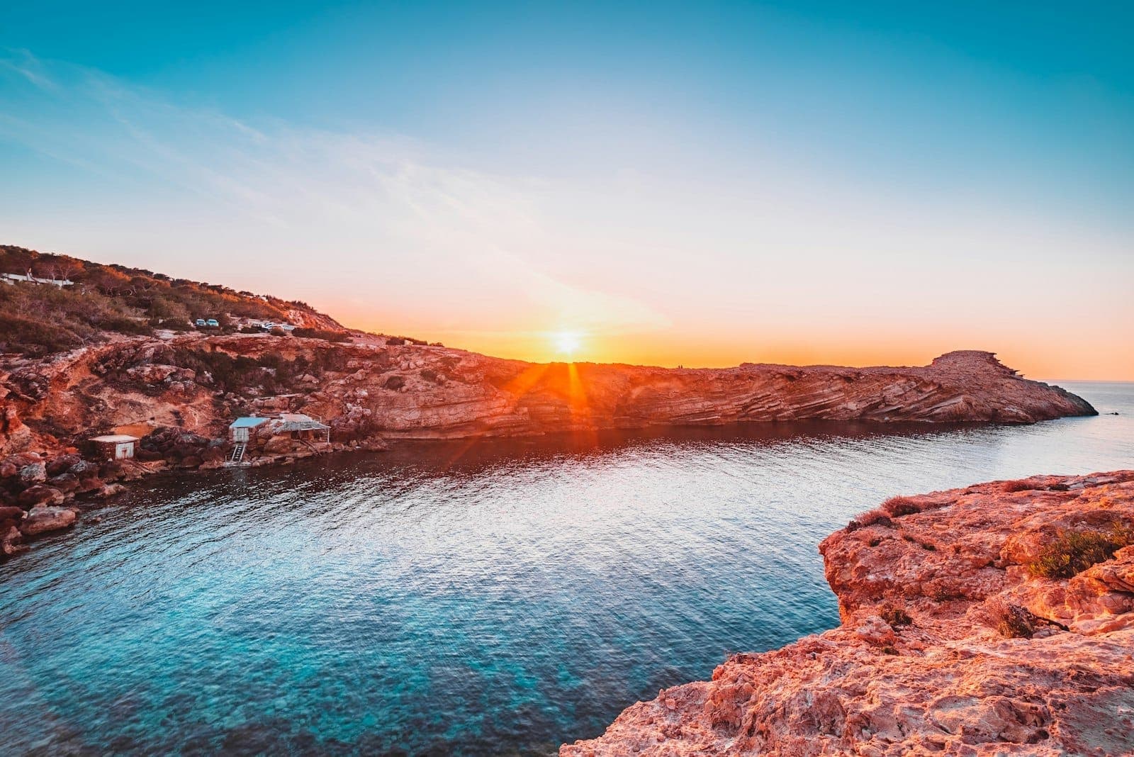 Rocky coastline and turquoise water at Ibiza’s Cala Comte beach, with the sun setting behind rugged cliffs under a clear sky.