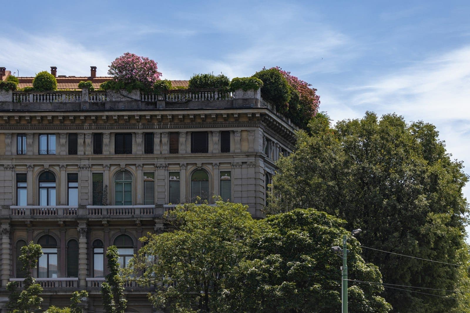 Elegantes historisches Gebäude in Mailand mit üppigen grünen Bäumen und rosa blühenden Blumen auf dem Dach, unter einem klaren blauen Frühlingshimmel.