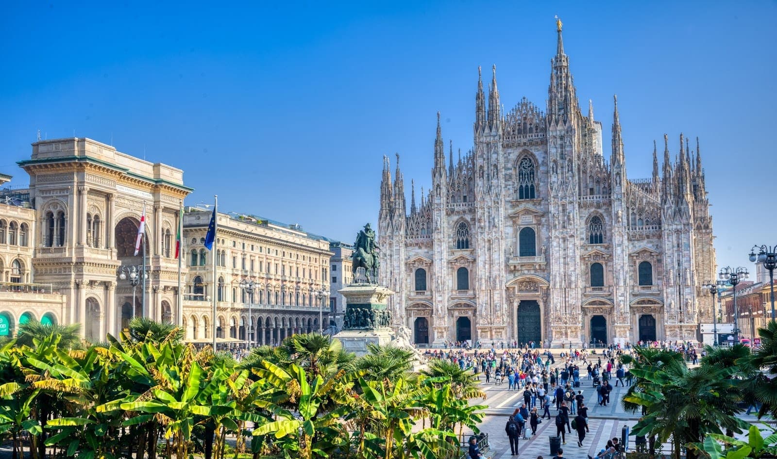 View of the Milan Cathedral and surrounding square bustling with people on a sunny day, with palm trees and clear blue sky in the foreground.