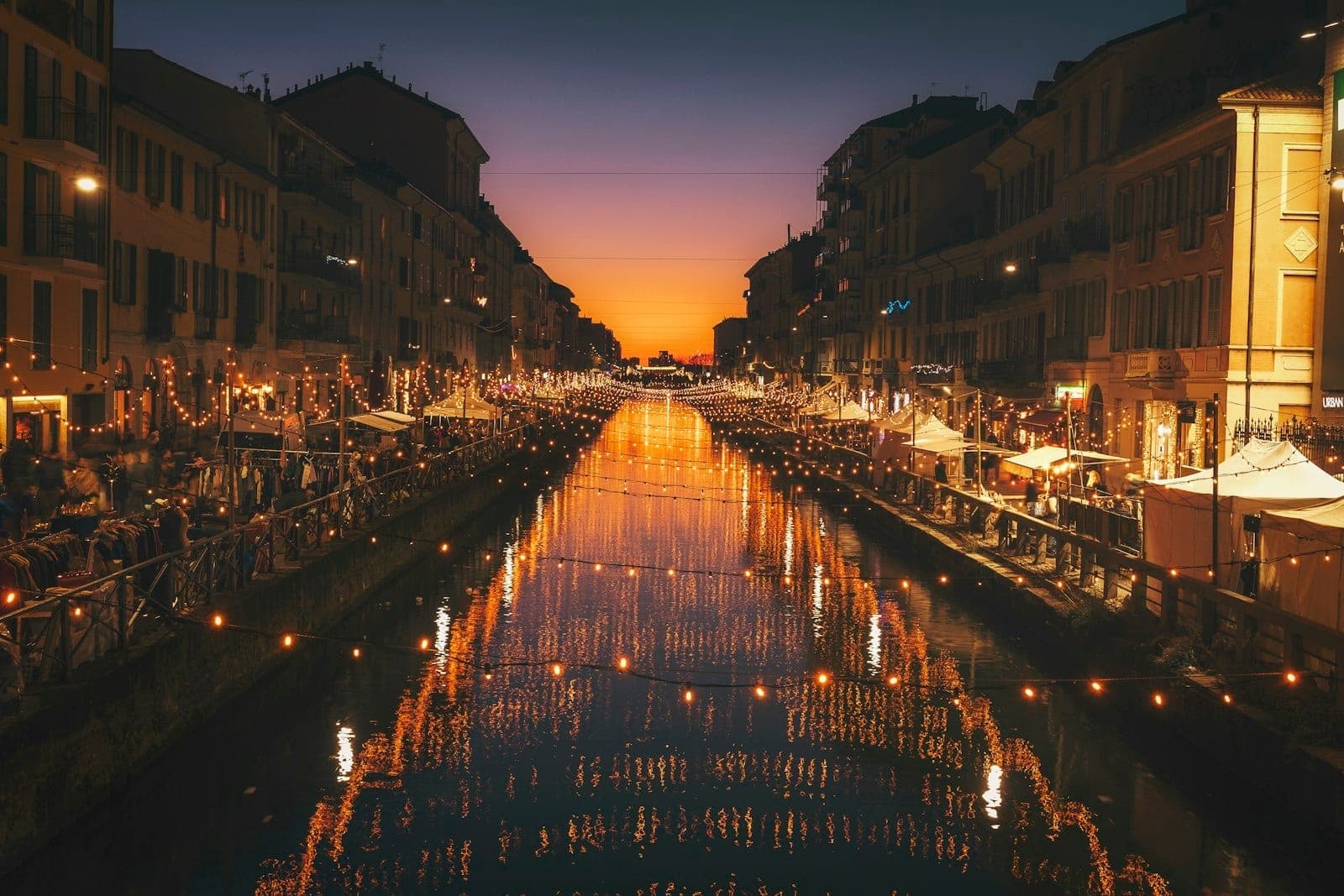 Der Navigli-Kanal in Mailand bei Sonnenuntergang, mit festlichen Lichtern, belebten Terrassen und einer lebhaften Sommerabendatmosphäre.
