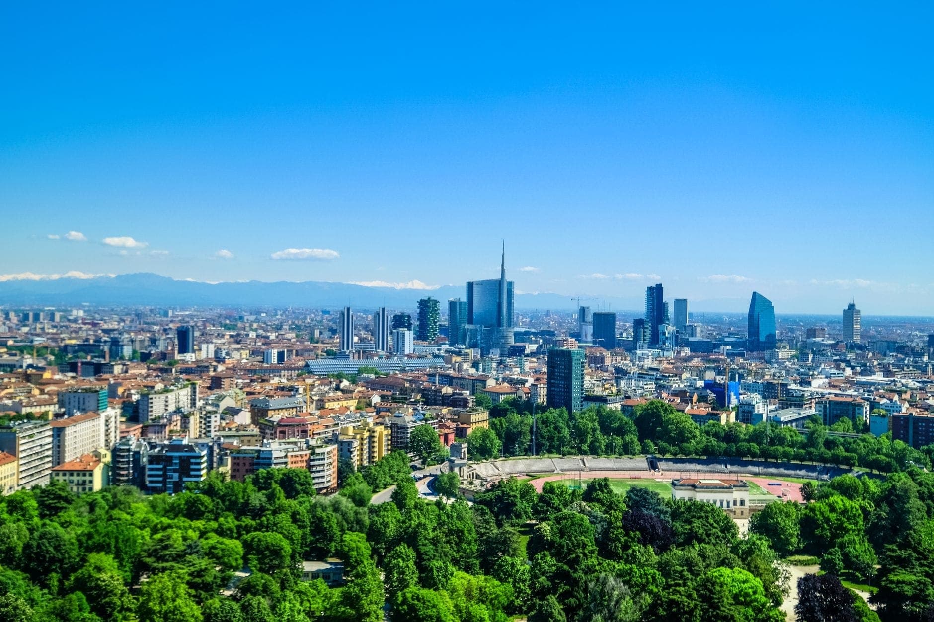 Panoramablick auf Mailand mit Stadtgebäuden, grünen Parks und den im Hintergrund sichtbaren Alpen unter einem sonnigen blauen Himmel.