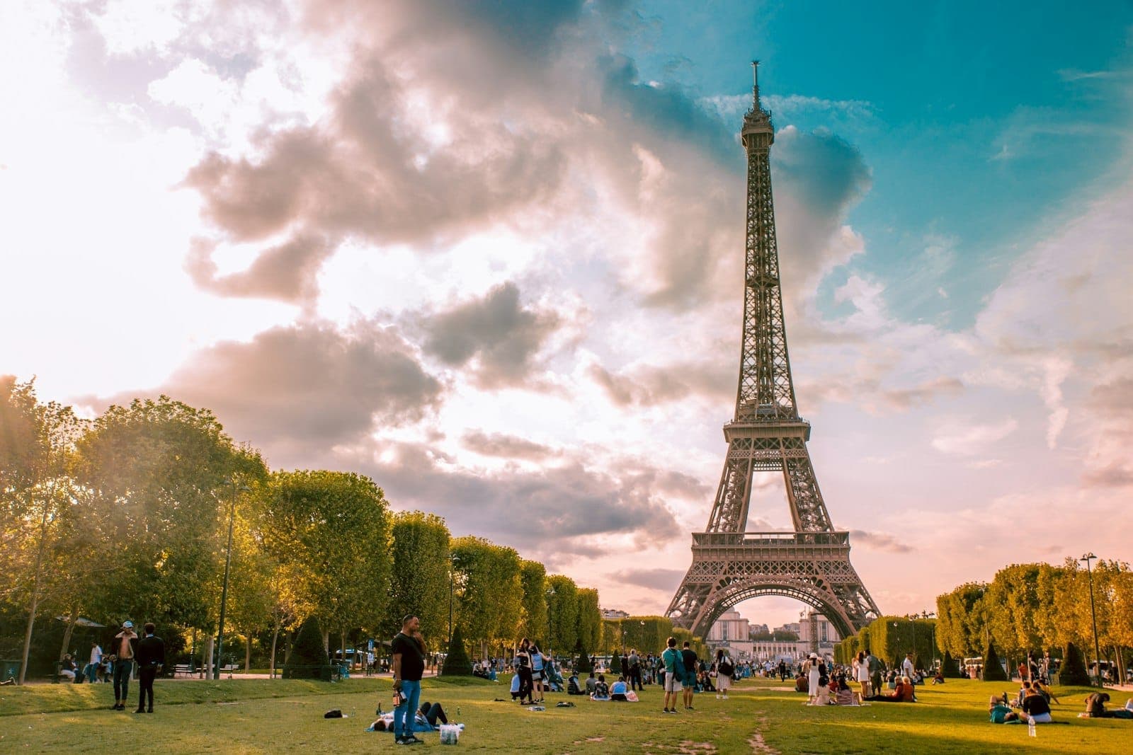 Tháp Eiffel nhìn từ Champ de Mars với những người đang thư giãn trong công viên dưới bầu trời có mây, gợi lên tinh thần du lịch và sự thay đổi theo mùa ở Paris.