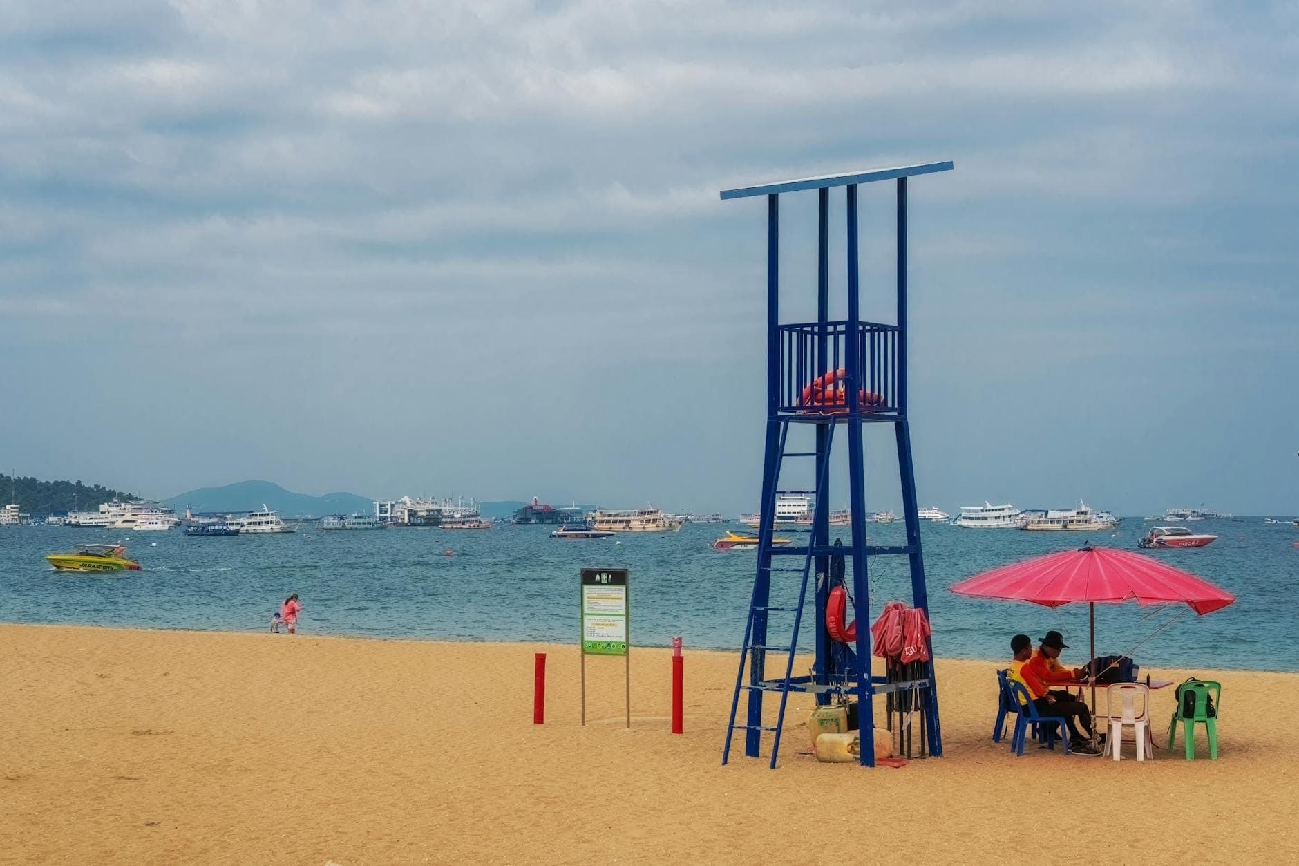 Sandy Pattaya beach with a blue lifeguard tower, pink umbrella, and cloudy skies, with boats anchored in the background.