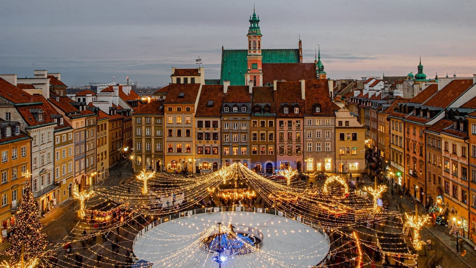 Rynek Starego Miasta w Warszawie zimą z świątecznymi lampkami bożonarodzeniowymi, lodowiskiem, kolorowymi historycznymi kamienicami i dużym kościołem w tle.