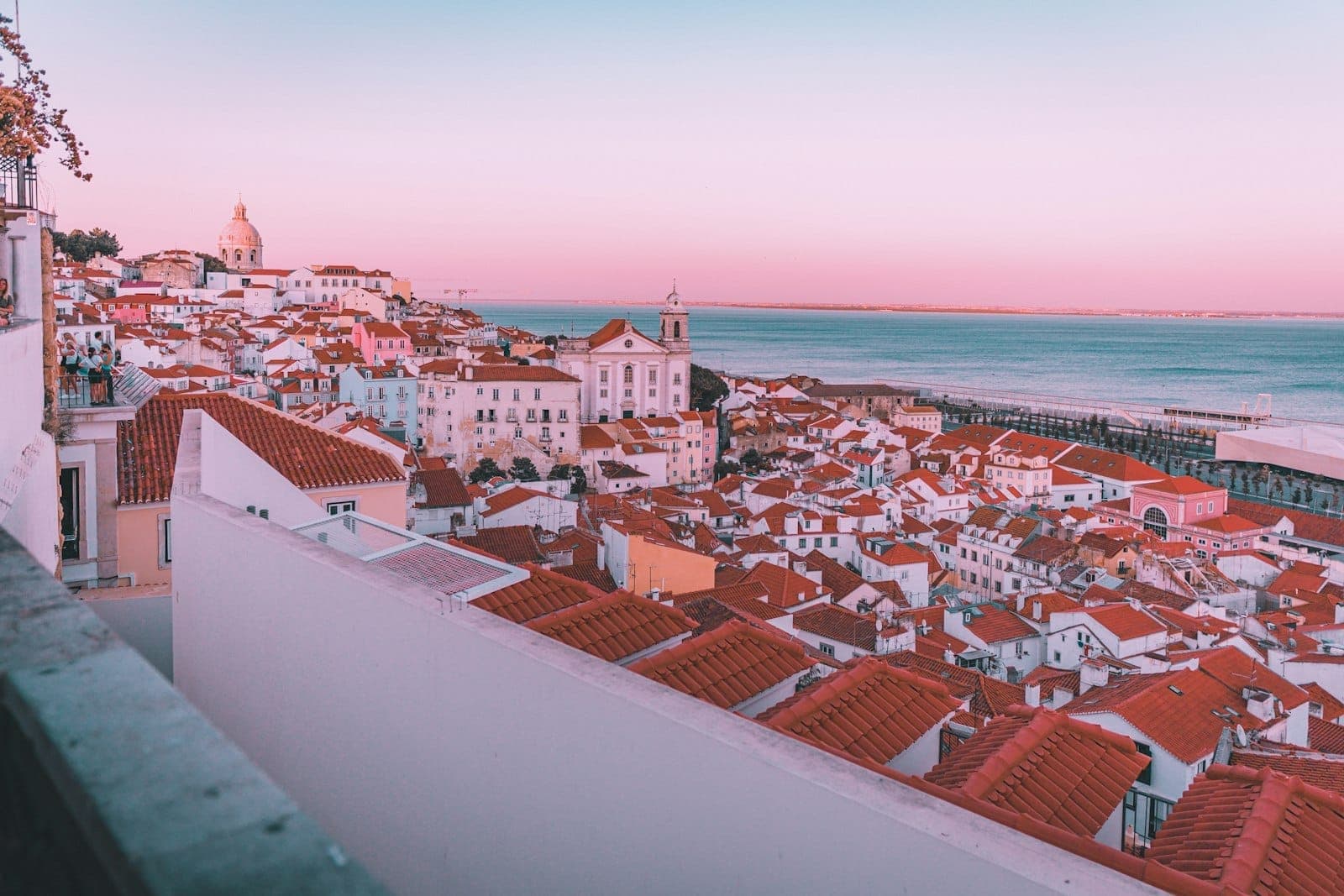 Toàn cảnh Lisbon với mái ngói đỏ rực và dòng sông Tagus trong ánh bình minh hay hoàng hôn, bầu trời hồng pastel và những tòa nhà lịch sử mang tính biểu tượng.
