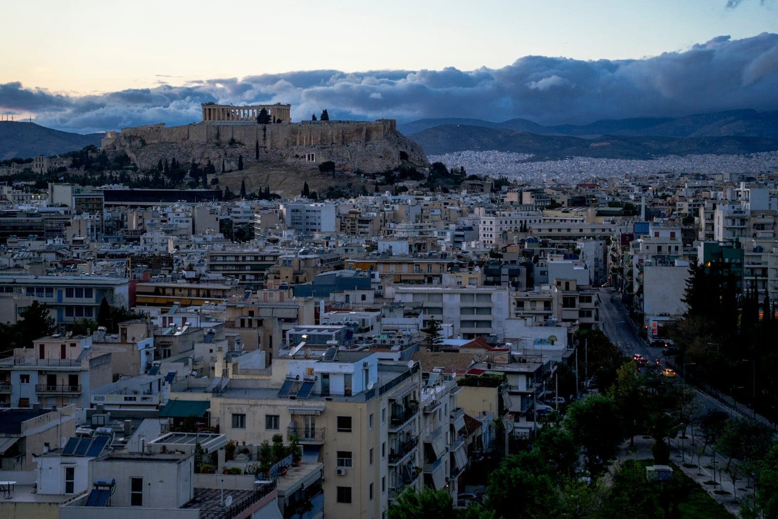 Panorama di Atene al tramonto con vedute dai tetti e l'Acropoli illuminata su una collina sullo sfondo, edifici urbani in primo piano.