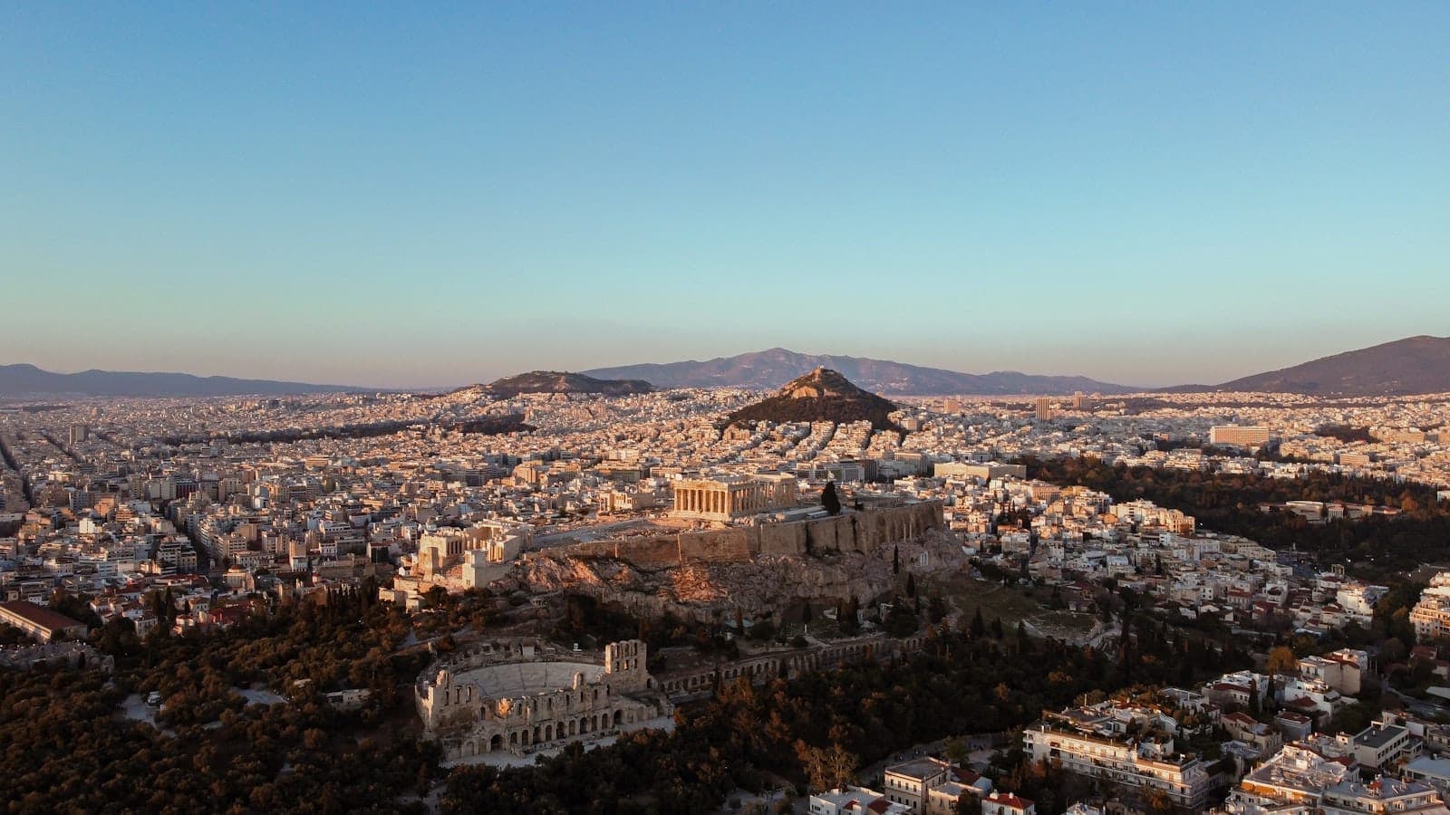 A sweeping panoramic view of Athens at sunset, featuring the Acropolis, Parthenon, cityscape, and distant hills under a clear sky.